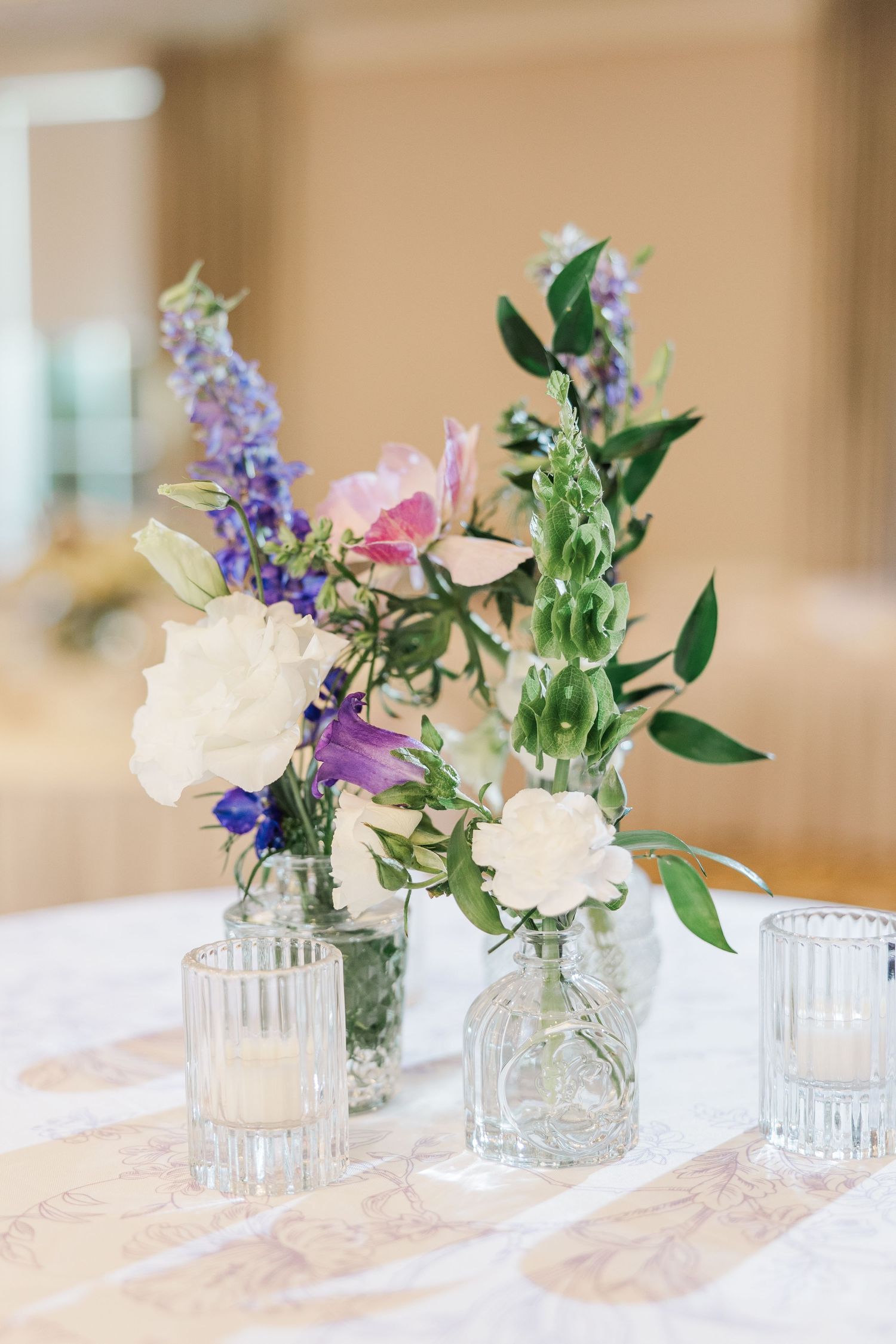 Small crystal vases with delicate purple and pink flowers arranged in a row on a white tablecloth.
