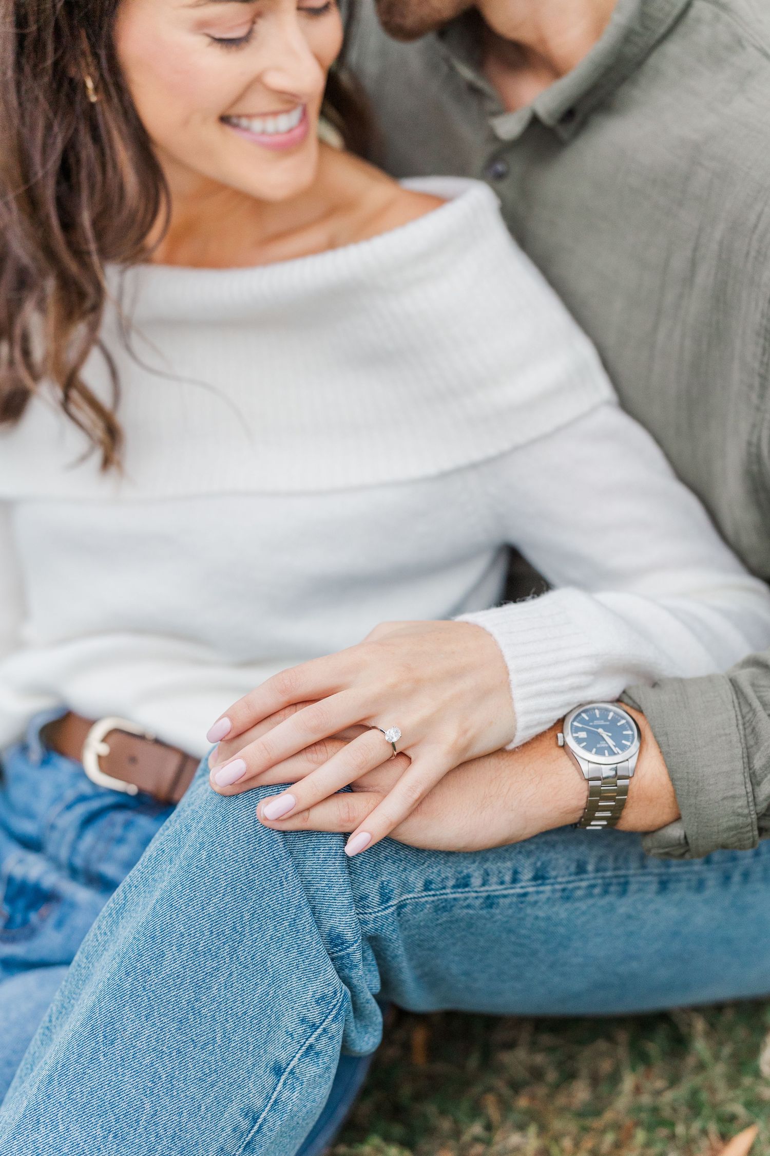 Close-up of couple showing engagement ring and watch while sitting together in casual denim and white sweater.
