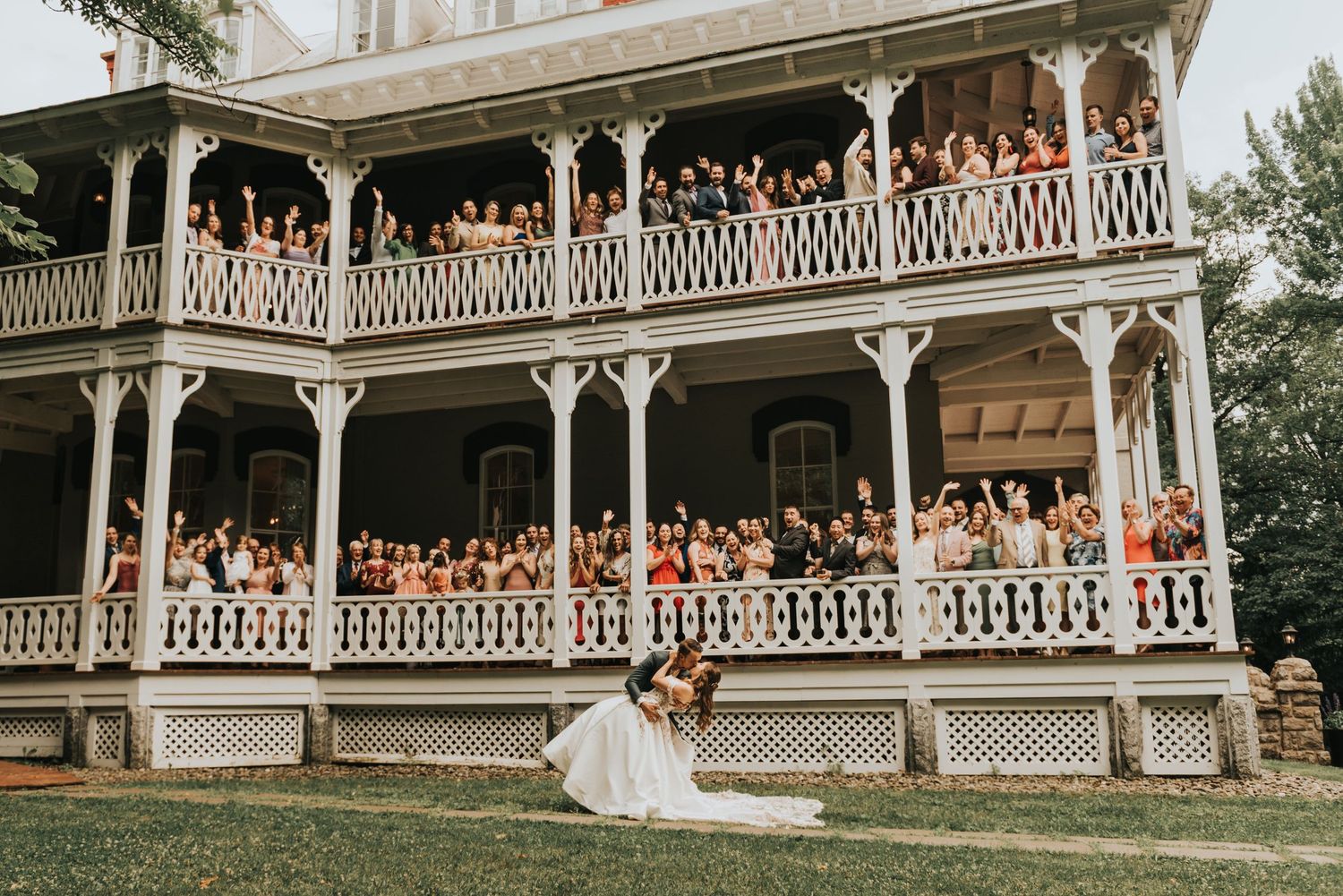 Wedding guests gather on a white ornate Victorian-style wraparound porch during an outdoor celebration.