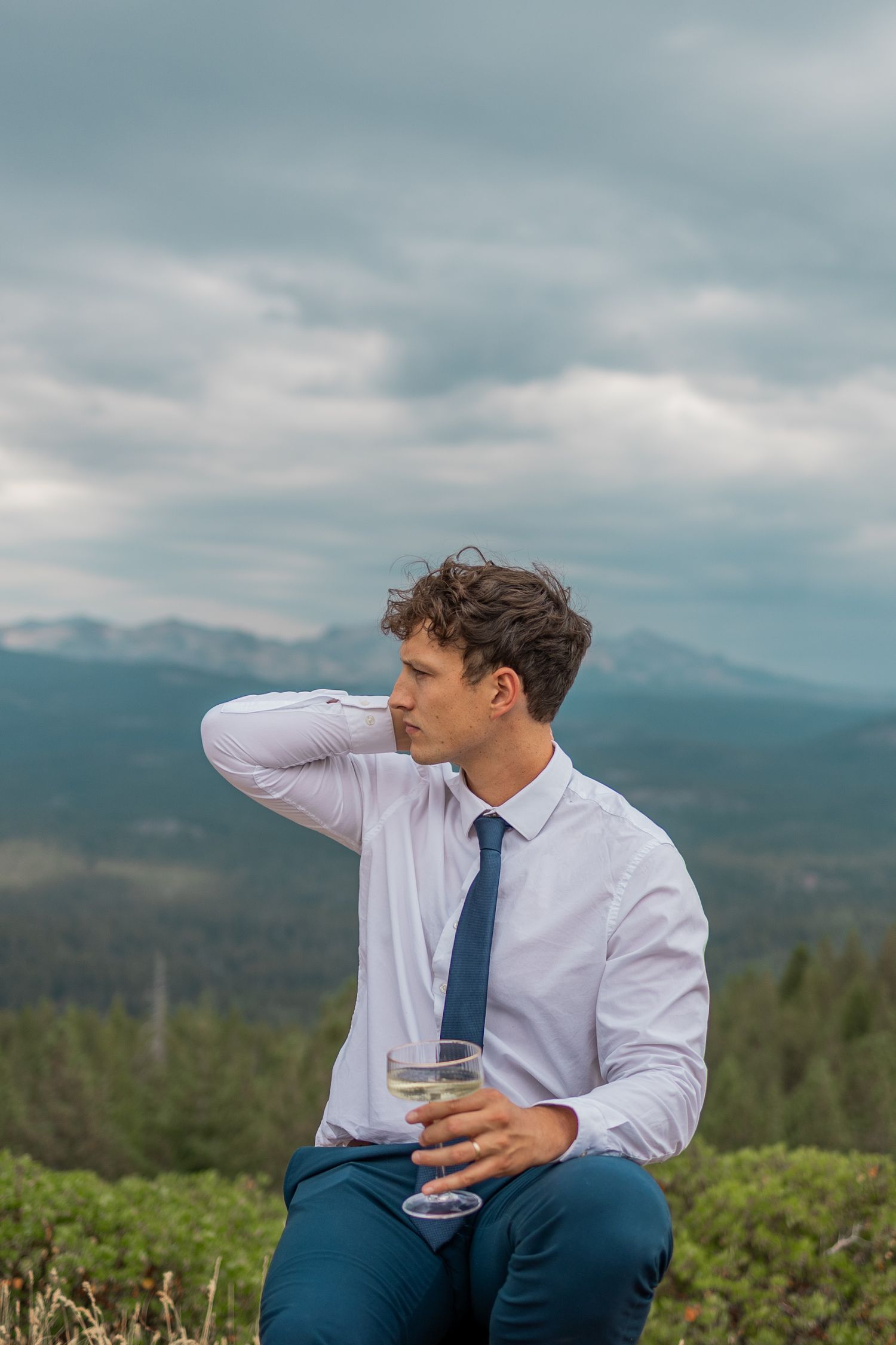 Person in business attire sits outdoors holding a drink while enjoying mountain views on a cloudy day.