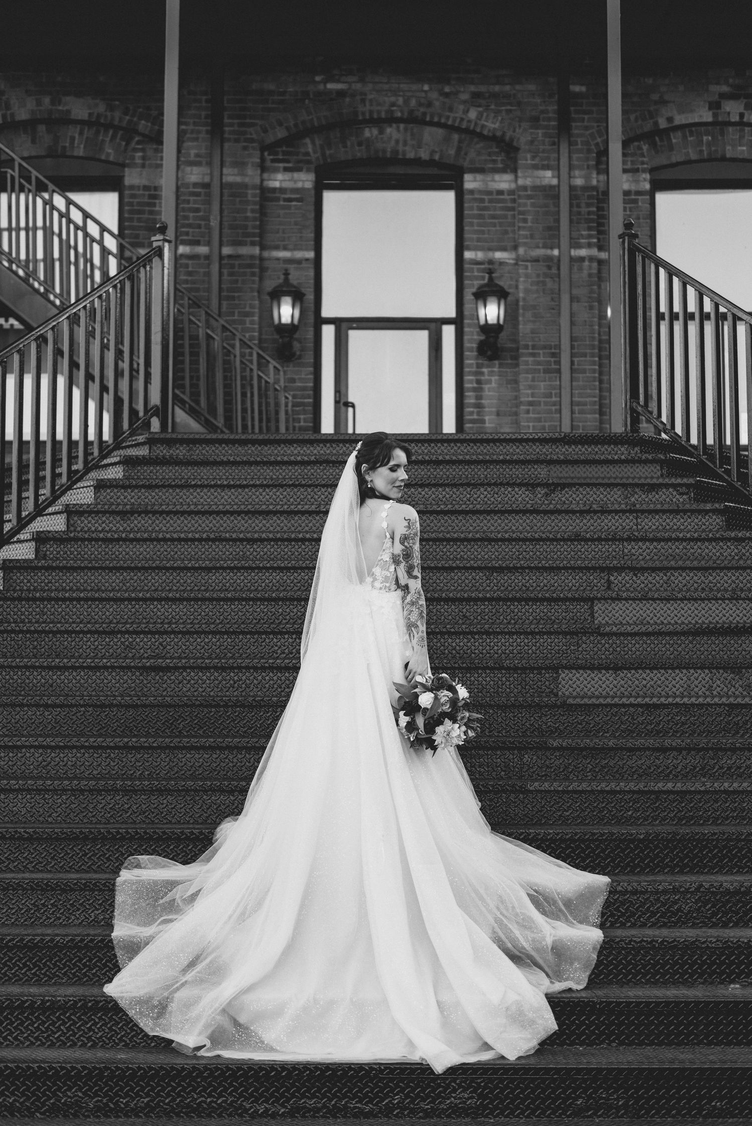 A bride in a flowing white wedding gown and veil poses gracefully on grand stone steps in a black and white photograph.
