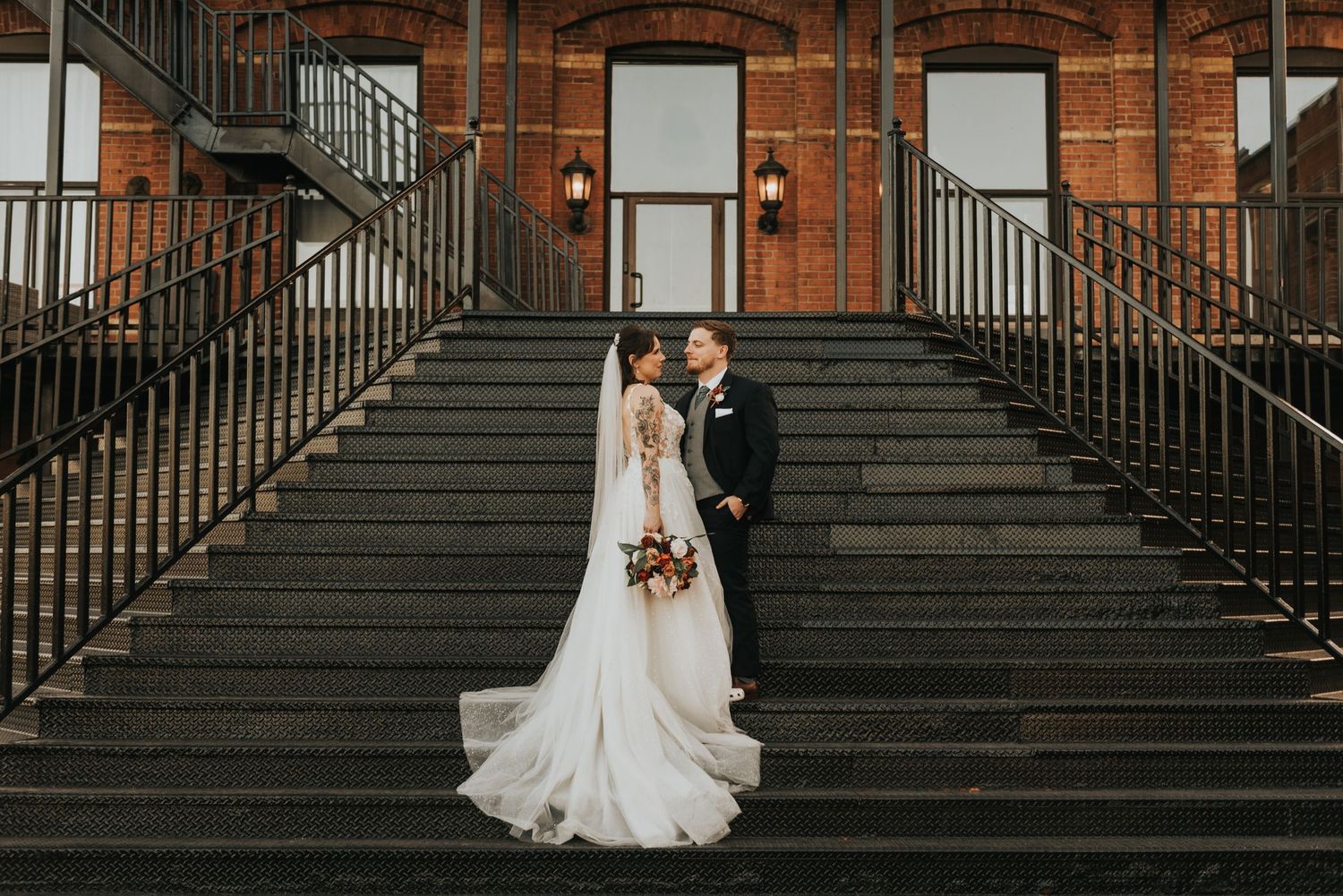 A romantic wedding couple poses on elegant black iron stairs against a red brick building backdrop.