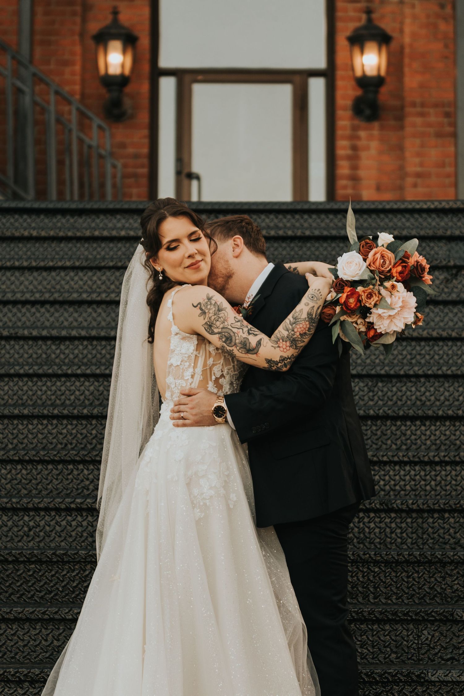 Intimate wedding portrait captured on black iron stairs with warm lantern lighting and brick architecture.