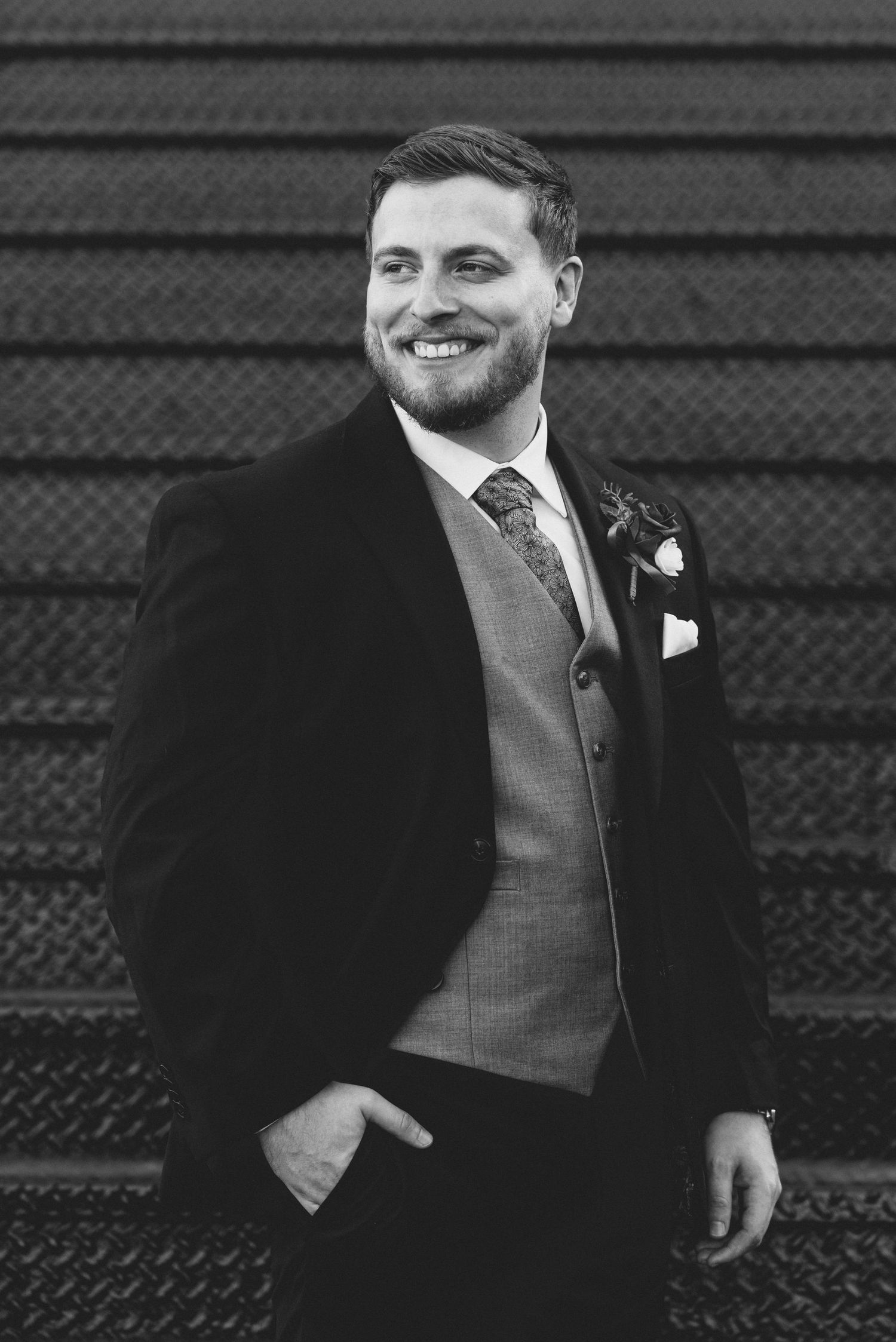 A groom in a dark suit and grey vest smiles warmly while standing against a black textured background.