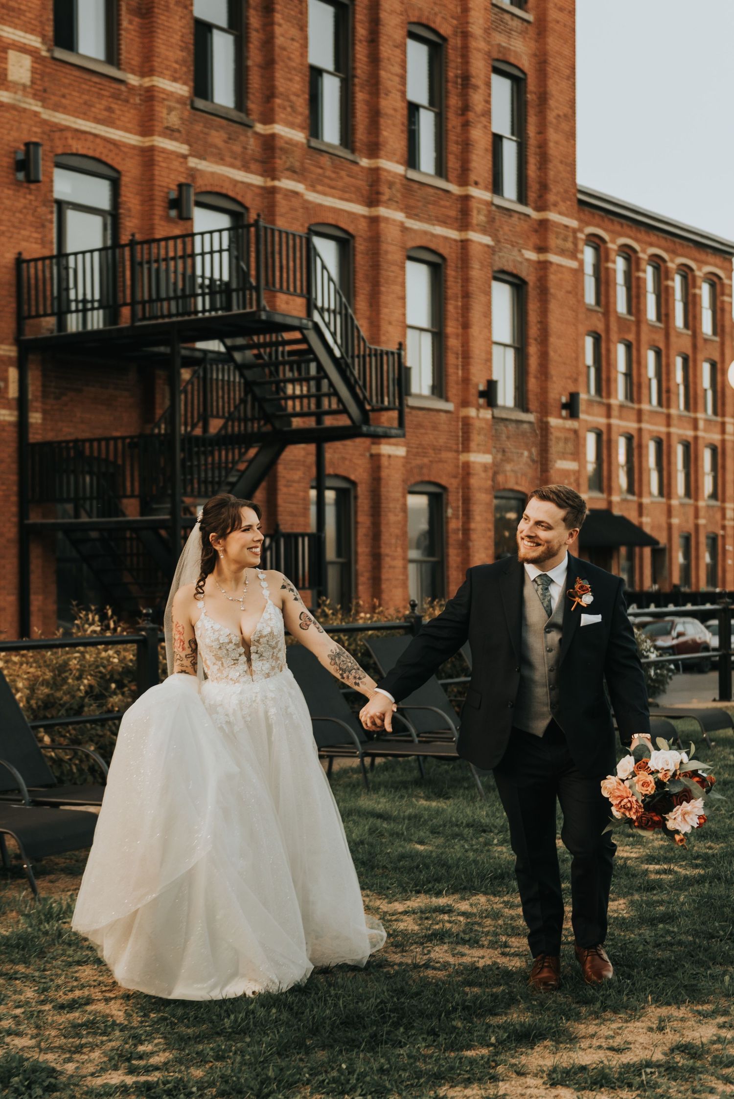 A joyful wedding couple shares candid moments against a brick building backdrop.