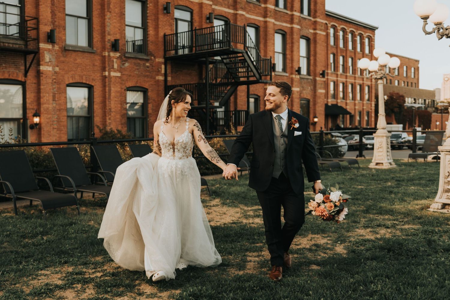 A couple in formal wedding attire walks through the grounds of a historic brick building during a photo session.