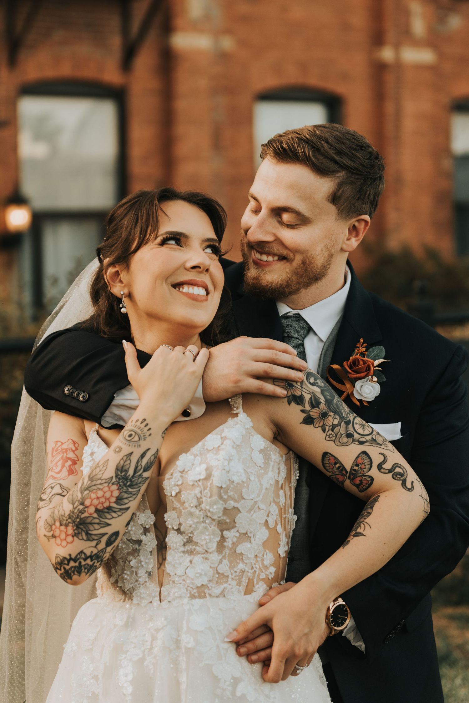 A tattooed bride in lace dress shares intimate moments with groom by historic brick building.