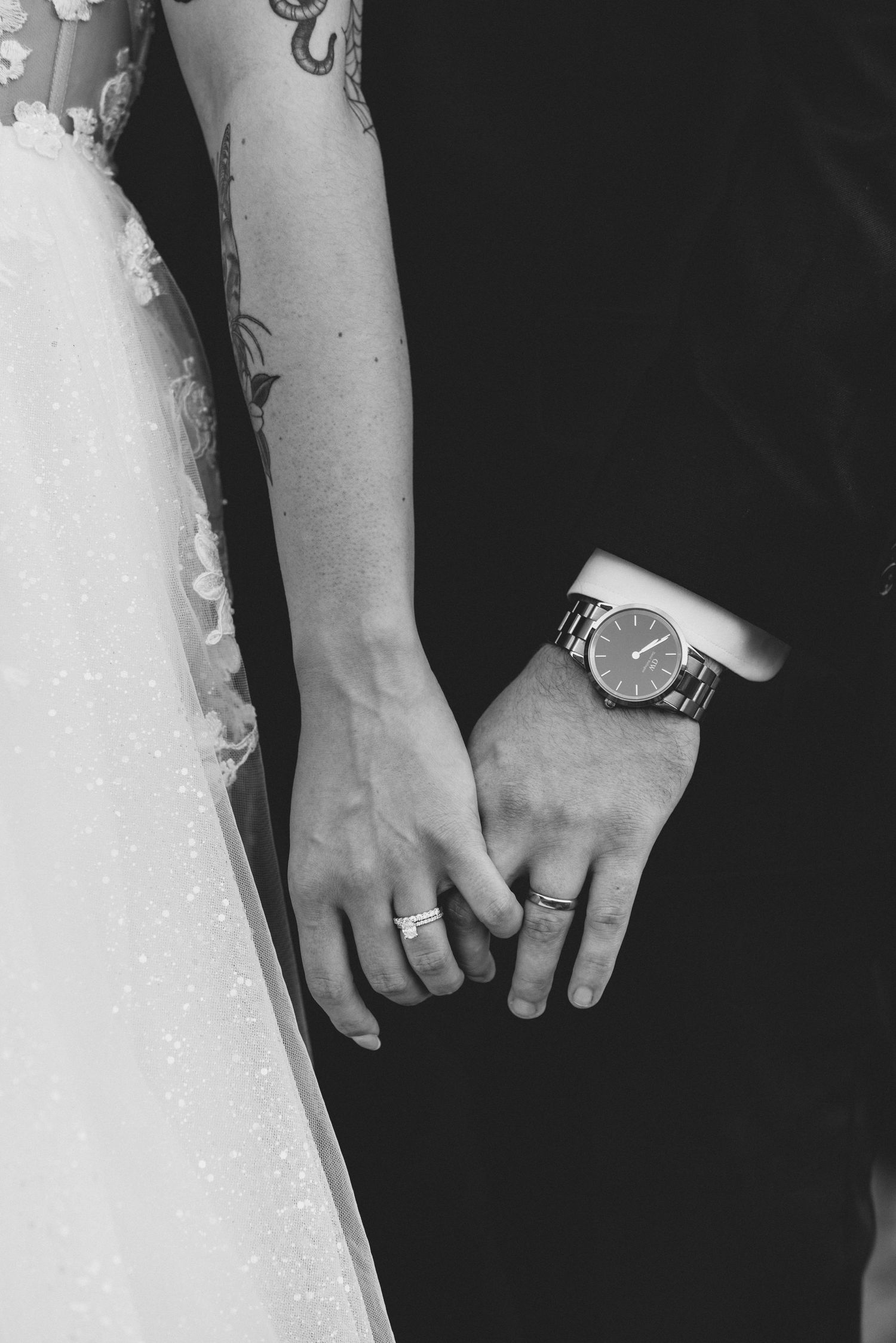 A black and white close-up of hands with wedding rings, showing the couple's joined hands against a dark background.
