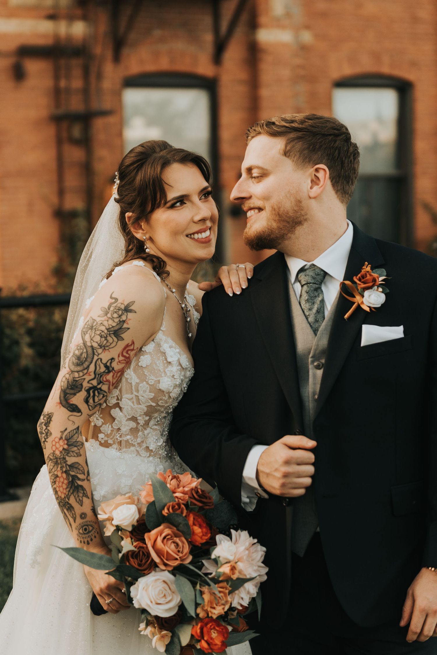 Newlyweds share romantic moments while holding coral and pink rose bouquet near brick walls.