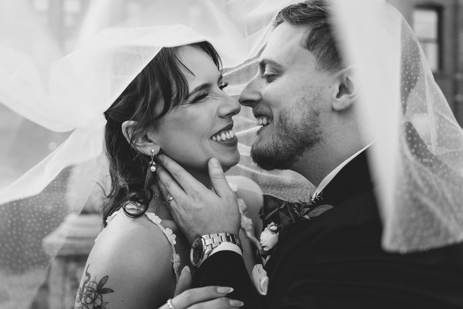 A romantic black and white wedding photo of a couple sharing a joyful moment under a flowing veil.