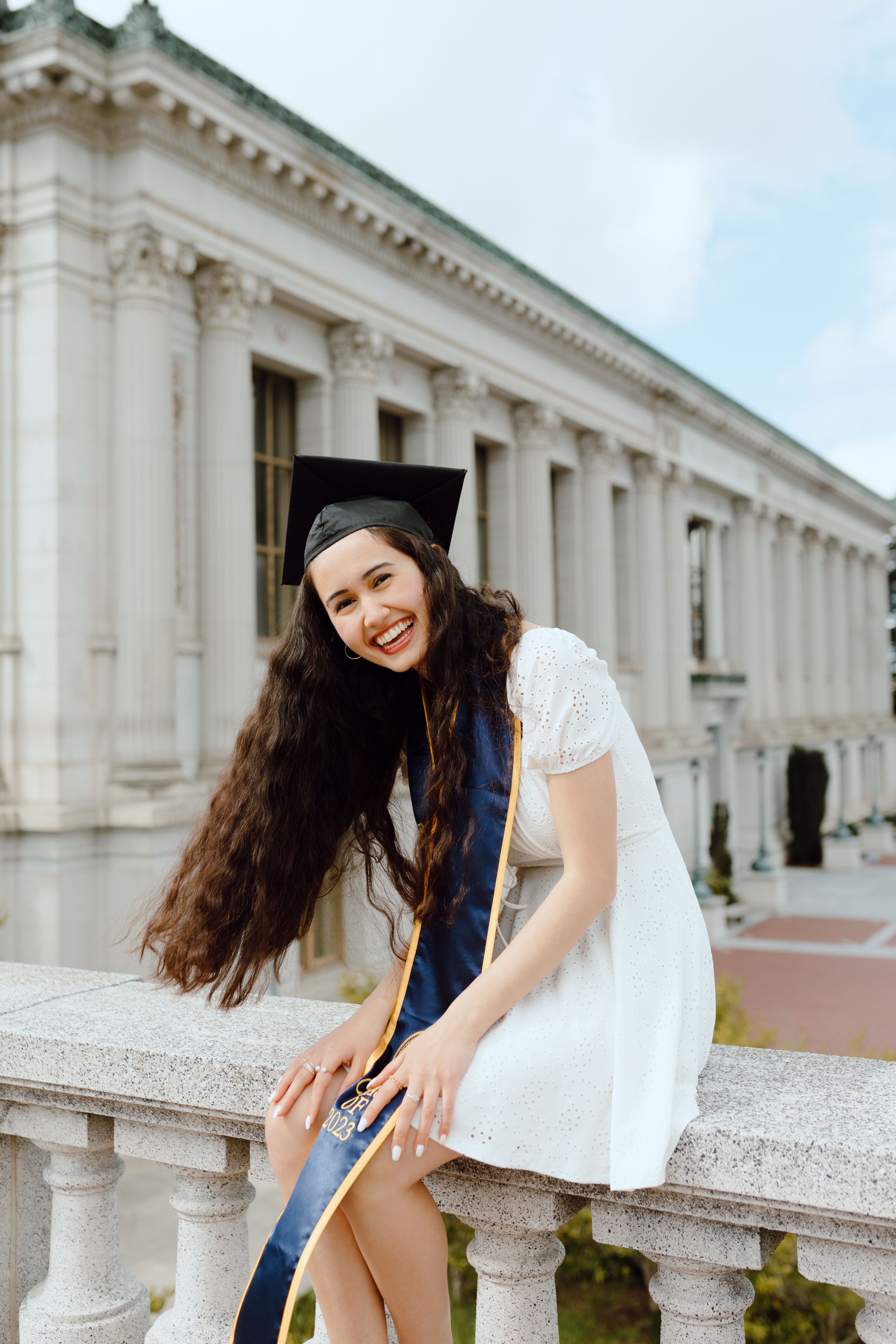 UC Berkeley graduate in white dress sits outside university building Doe Library for her grad photoshoot.