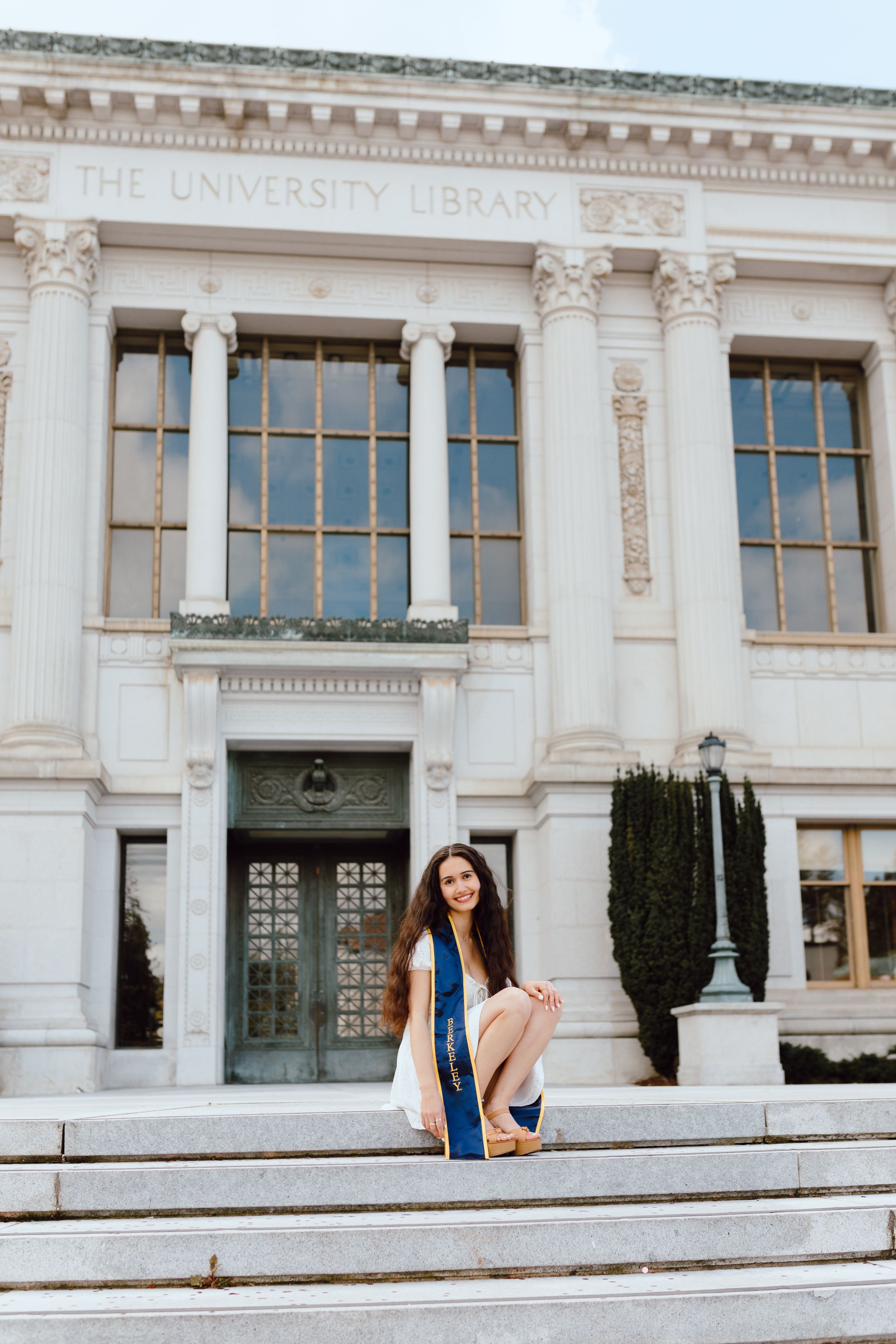 UC Berkeley graduate poses outside one of the campus's beautiful buildings (Doe Library) for her grad photoshoot. She wears a grad sash.