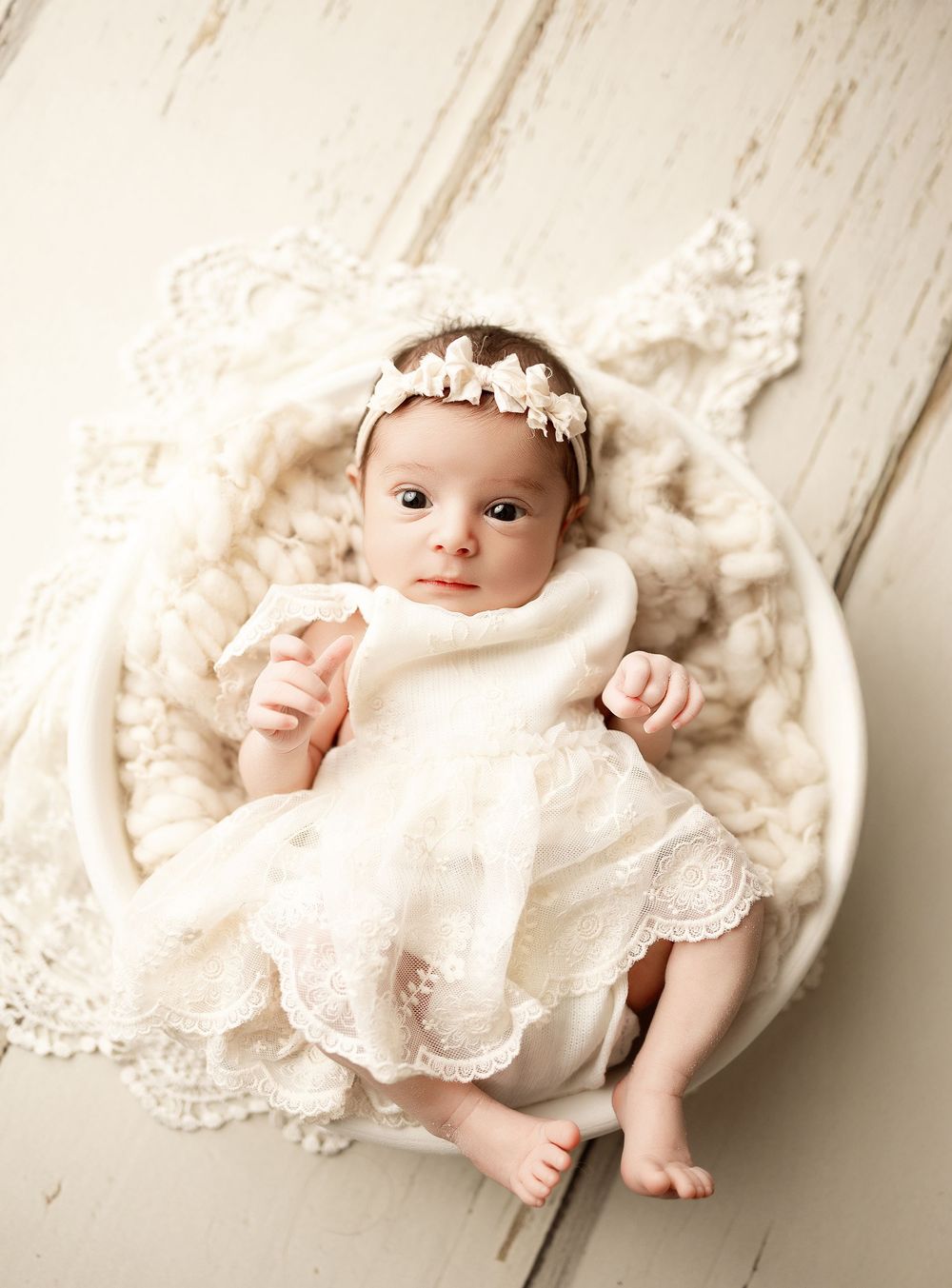 Newborn baby wearing white lace dress lying on cream-colored blanket in artistic photography series.