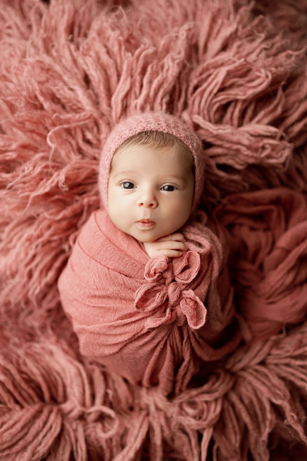 A newborn baby wrapped in pink swaddle cloth lying on a pink fluffy textured blanket in a professional photo shoot setting.