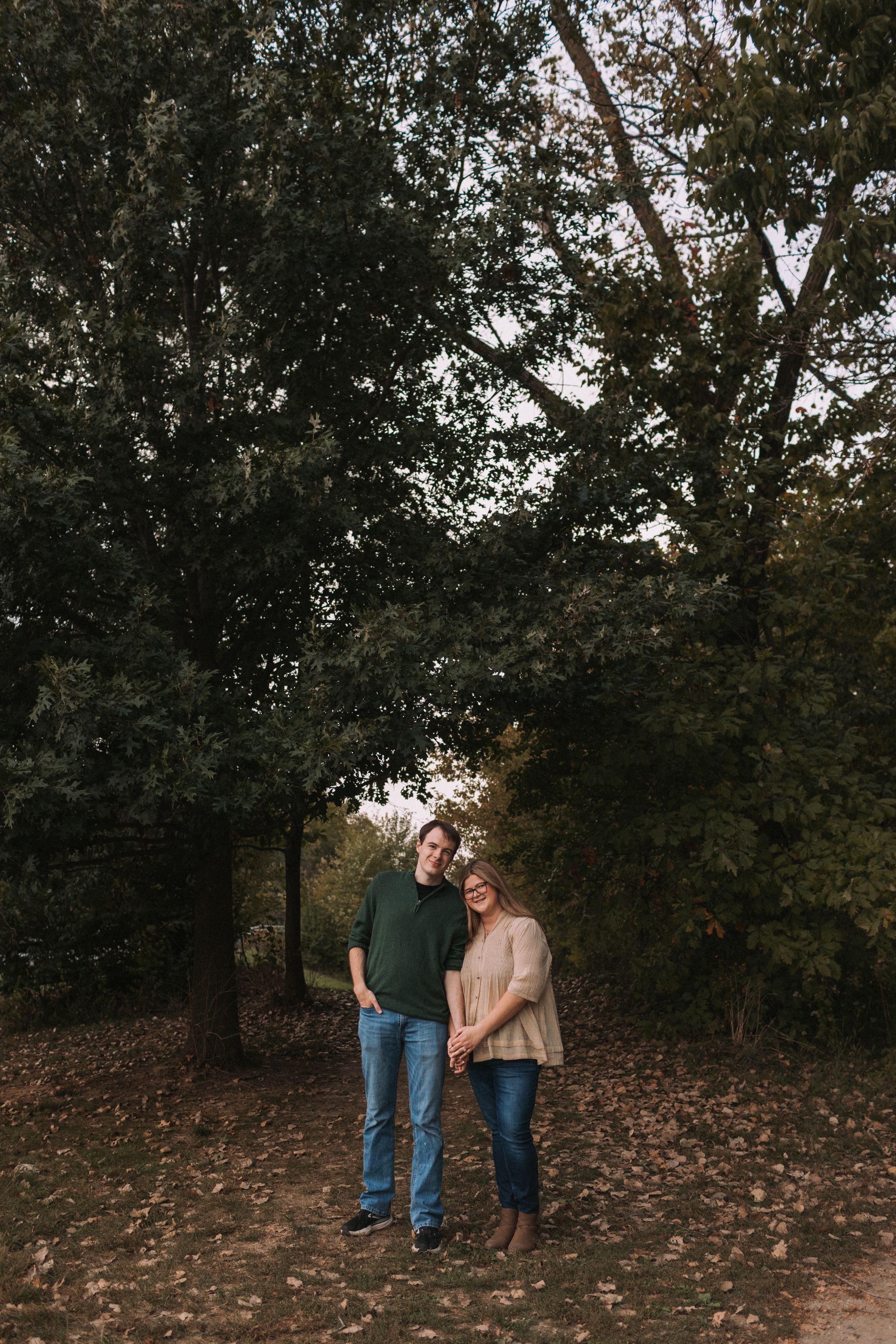 Couple stands together on fallen autumn leaves under tree canopy.