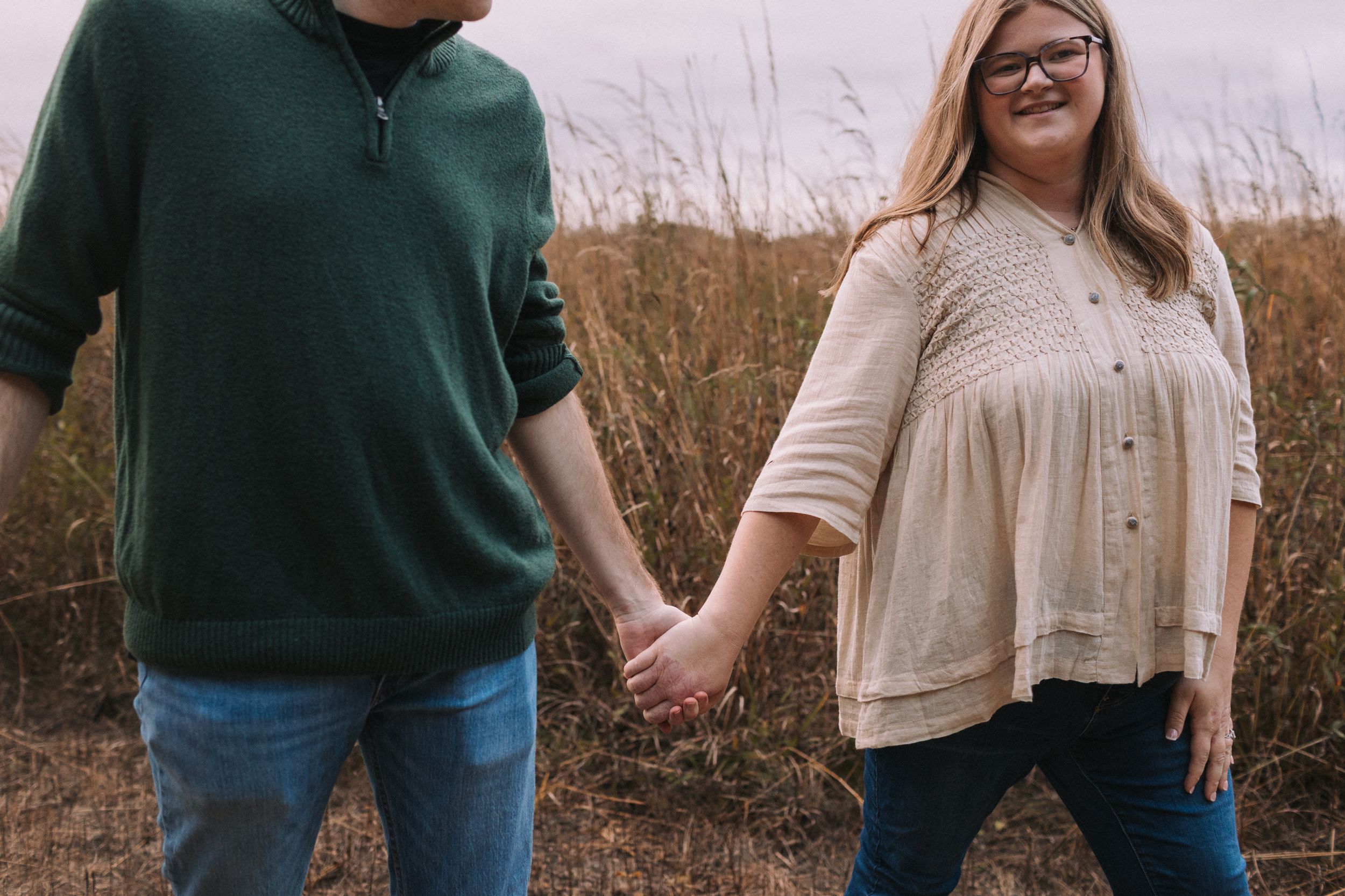 A playful couple in casual attire poses together in a field on an overcast day.