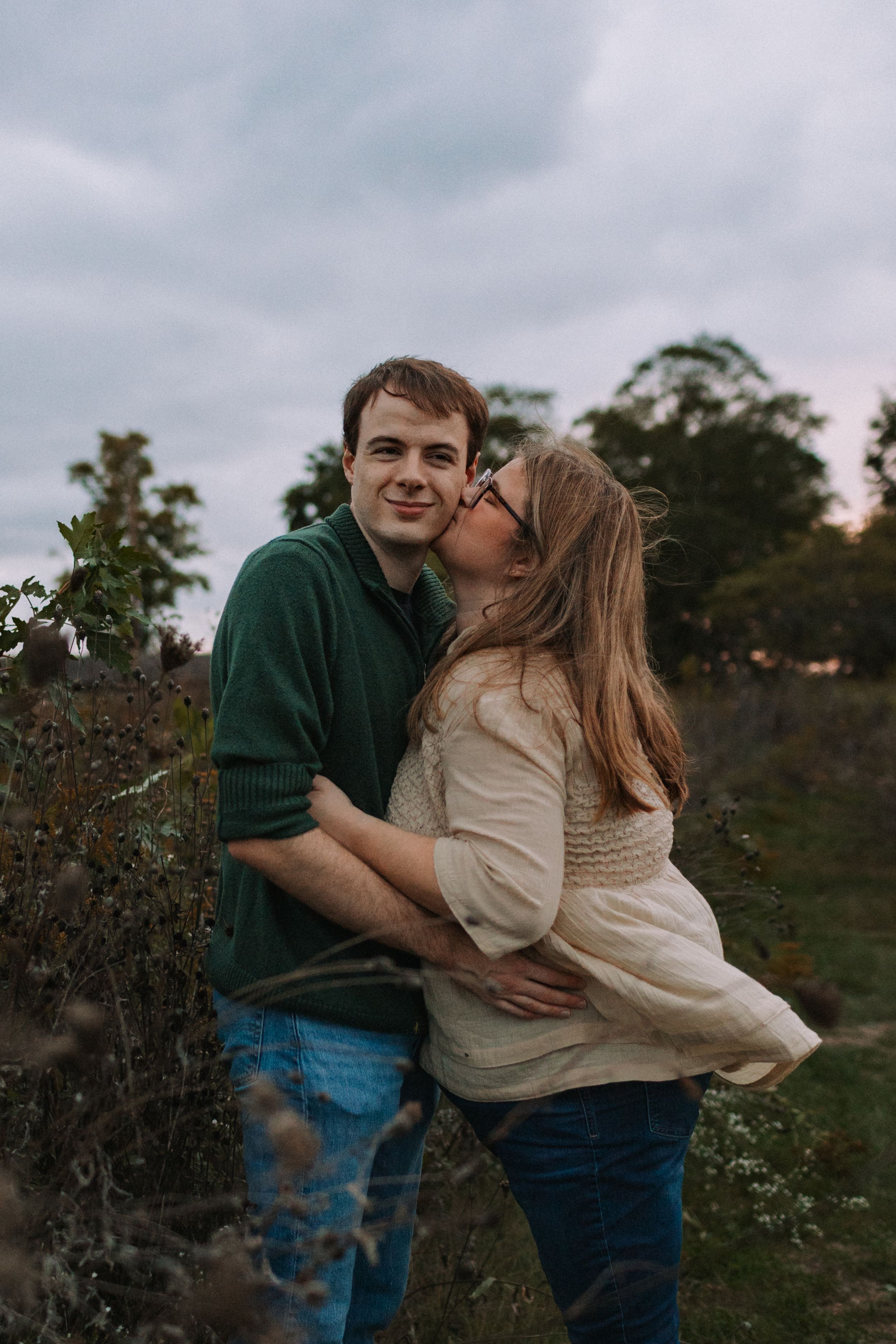 A playful couple embraces in a field during an overcast afternoon.