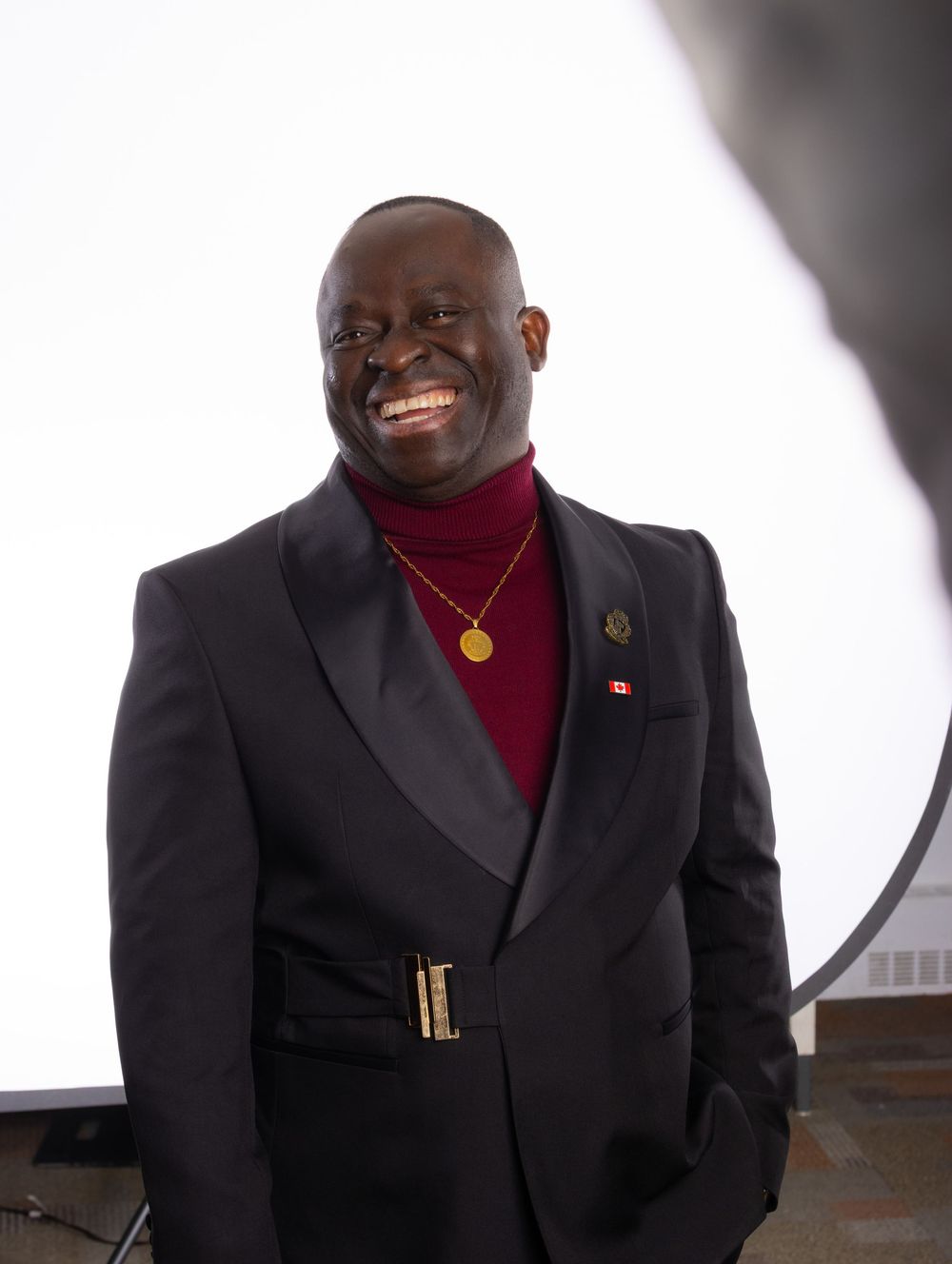 A person wearing a black suit jacket and burgundy turtleneck smiles warmly against a white background.