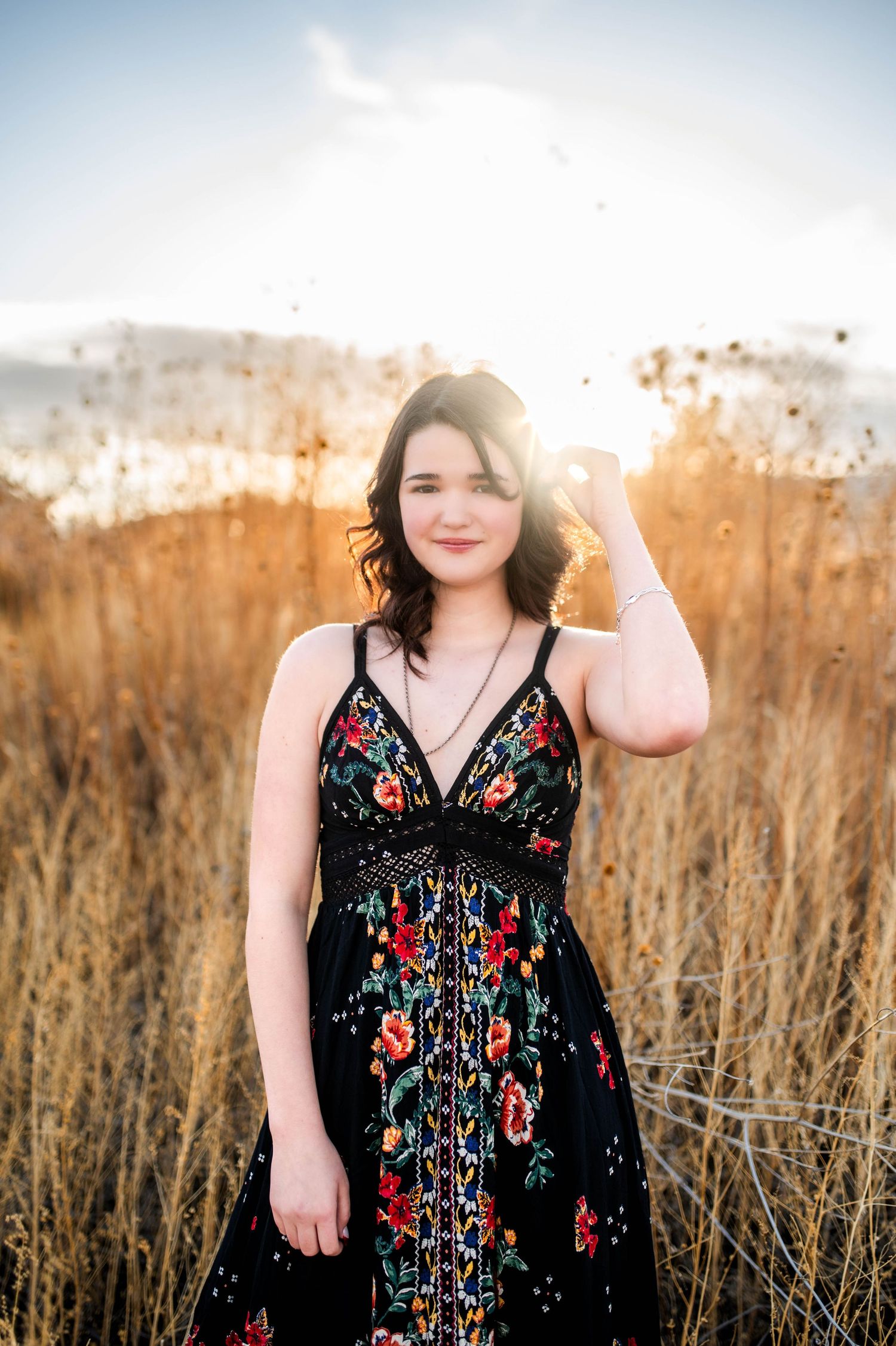 Young woman in black floral maxi dress standing in golden field at sunset with hand raised to hair.