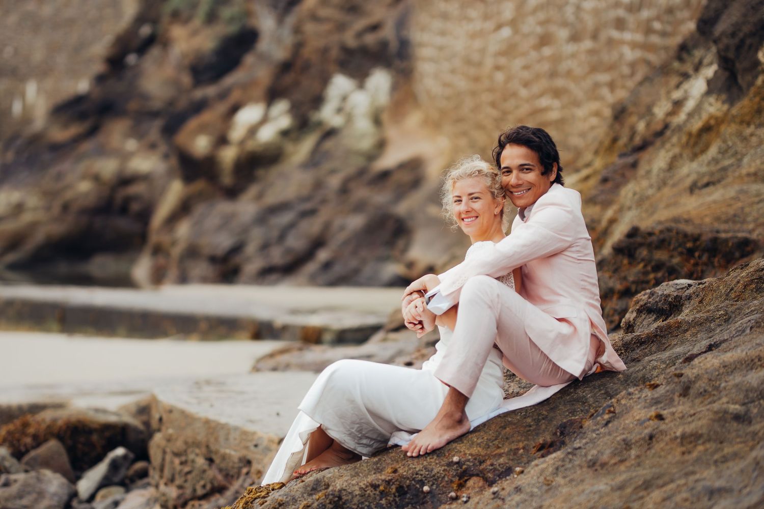 Two women in light casual outfits sitting together on rocky coastal cliffs smiling warmly.