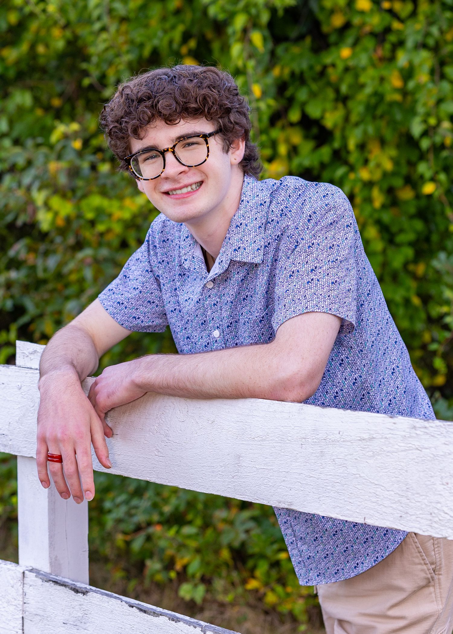 High school senior in a blue floral shirt and khaki pants leans casually on a white fence post in an outdoor garden setting.