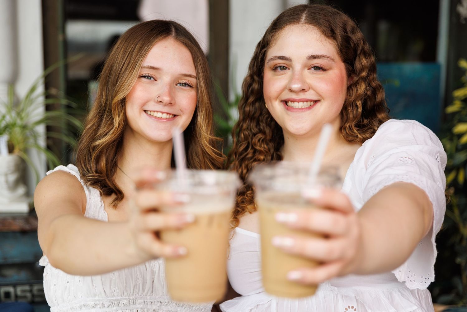Two friends in white tops smile while holding up iced coffee drinks for a toast at a cafe.