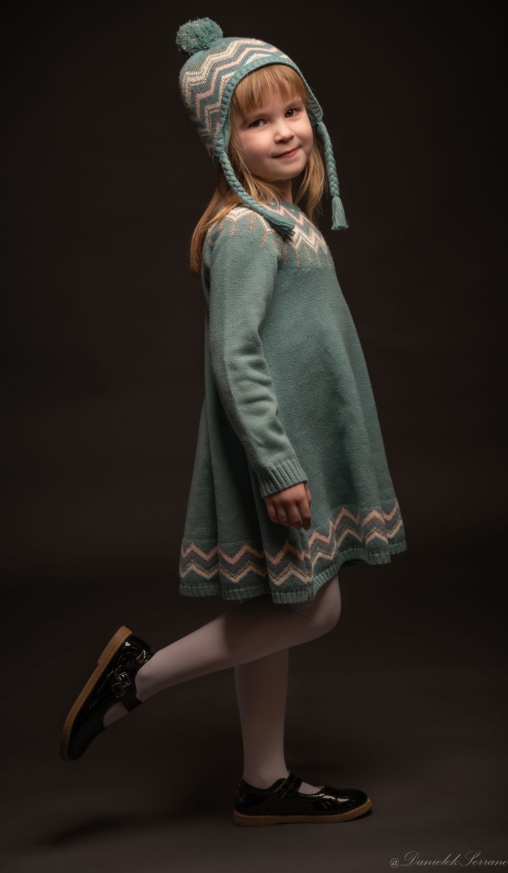 Little girl in teal knit dress and matching pom-pom hat posing against dark background.