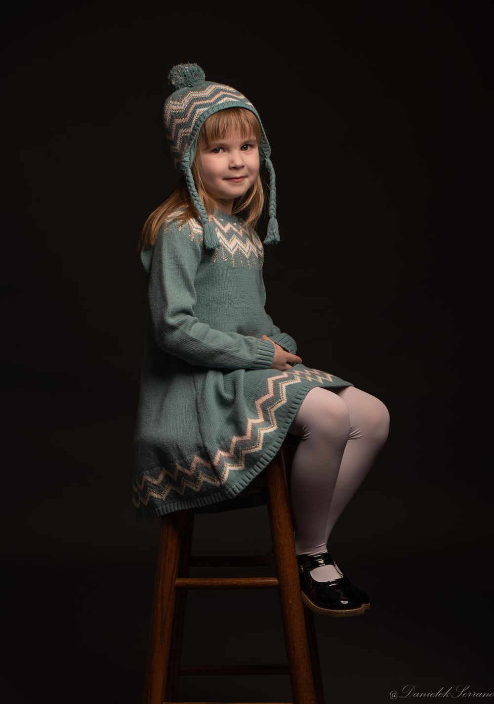 Young girl in teal Fair Isle knit dress and hat sitting on wooden stool.