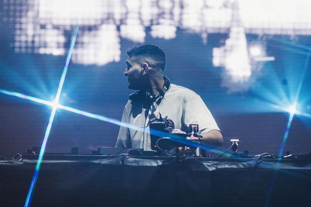 A performer in dark clothing crouches on stage against a backdrop of dramatic blue lighting and fog effects.