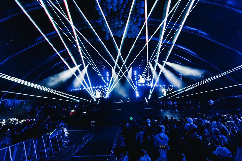 Concert stage illuminated with dramatic blue lighting and laser beams creating dynamic visual effects during performance.