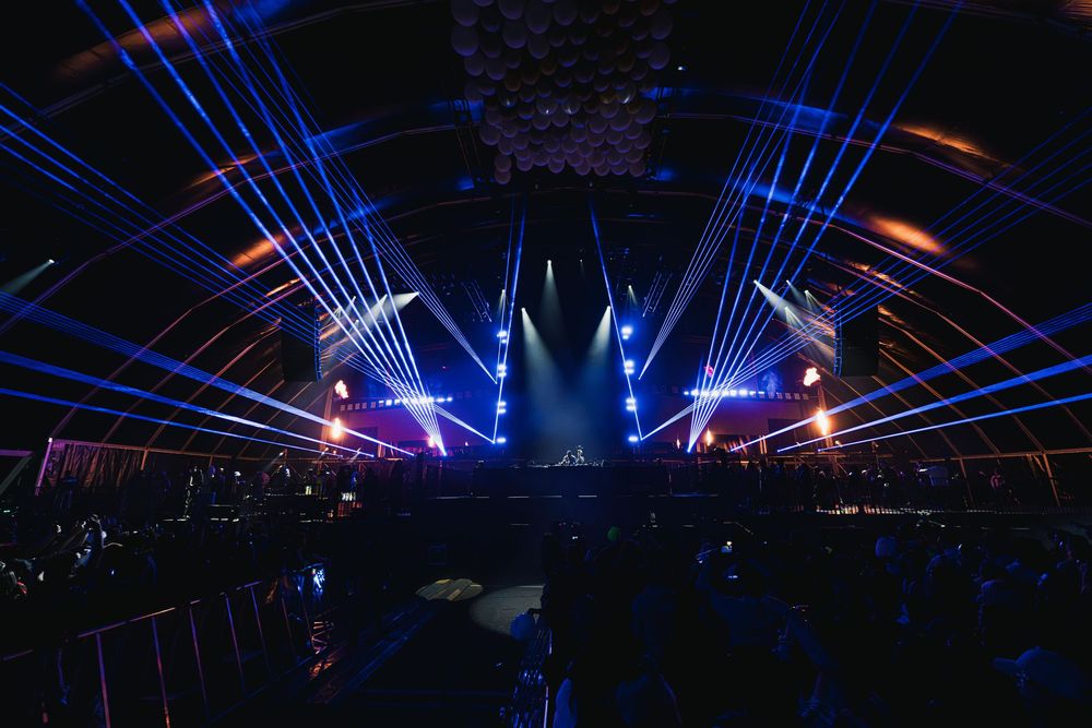 Spectacular concert stage with dramatic blue laser light beams and colorful lighting effects illuminating a dark arena.