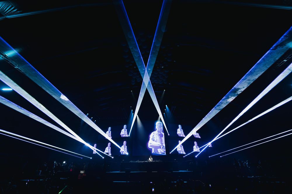 Dramatic stage performance with blue laser light beams creating geometric patterns against a dark background.