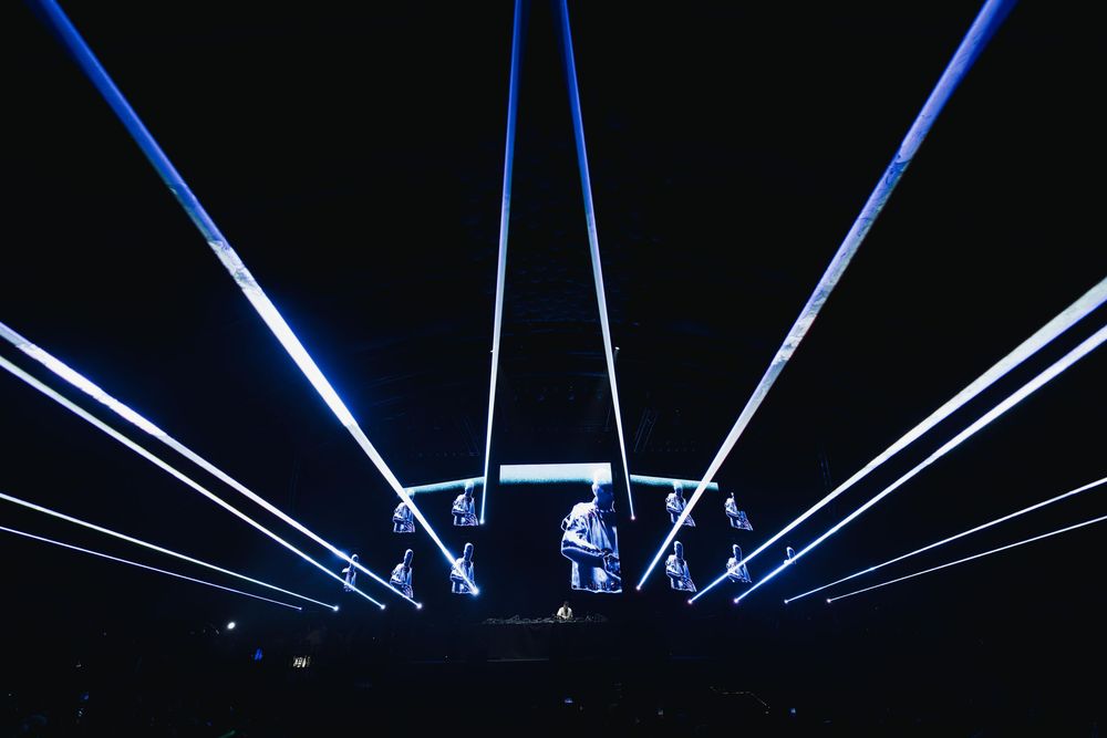 Bright blue laser beams illuminate a dark stage during a dramatic concert performance.