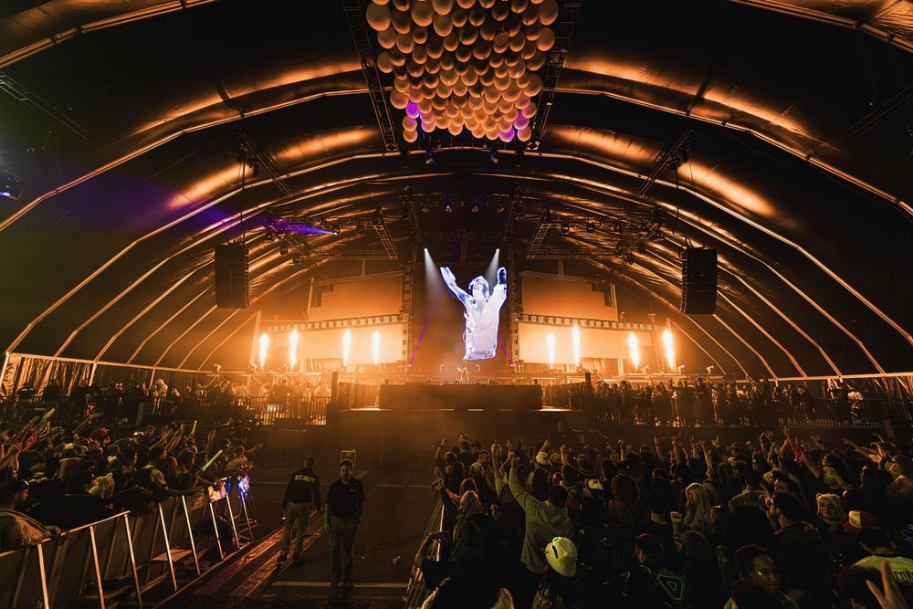 Concert crowd silhouetted against glowing orange stage lights in an arched venue during a live performance.