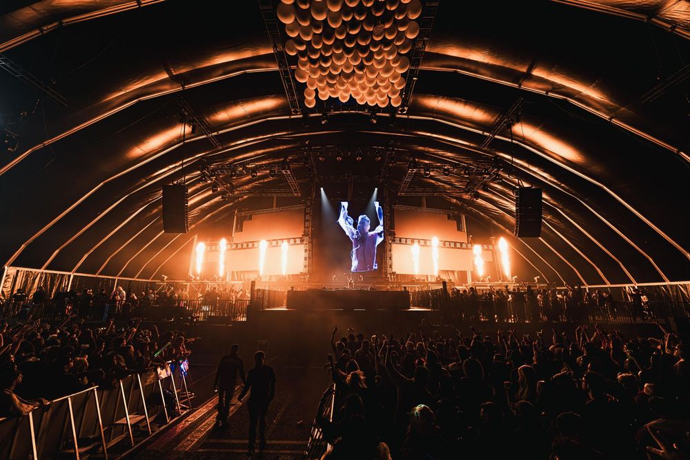 Concert crowd in a dark arena with dramatic orange lighting and illuminated stage design creating an atmospheric ambiance.