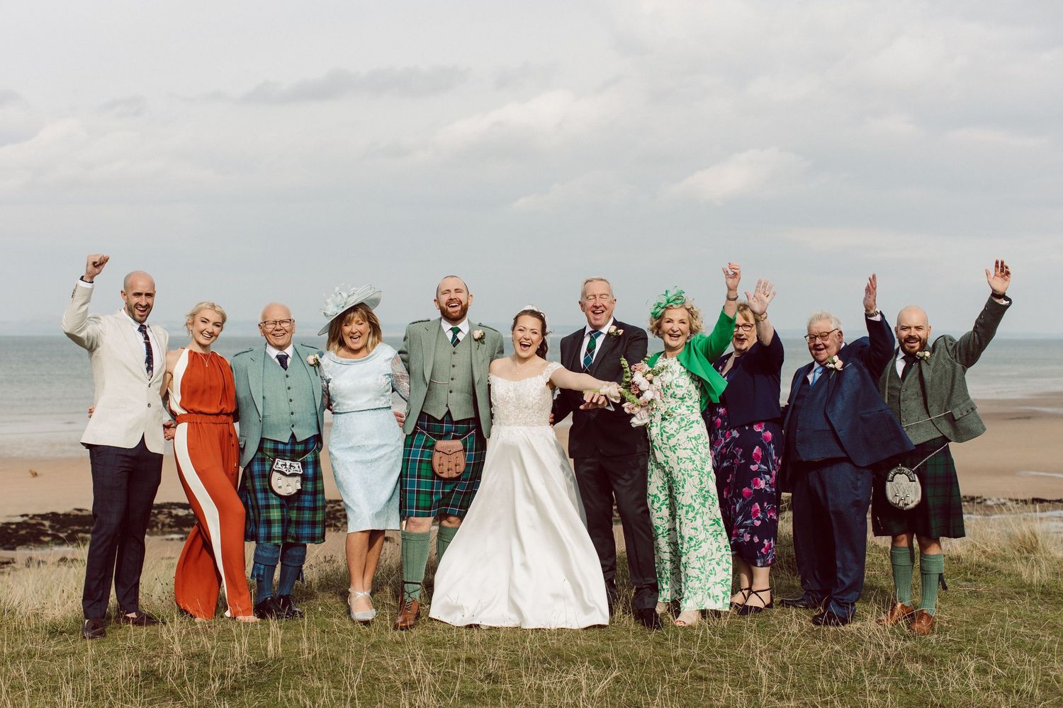 Wedding party jumping together on a grassy hill against a cloudy sky in a fun group photo sequence.