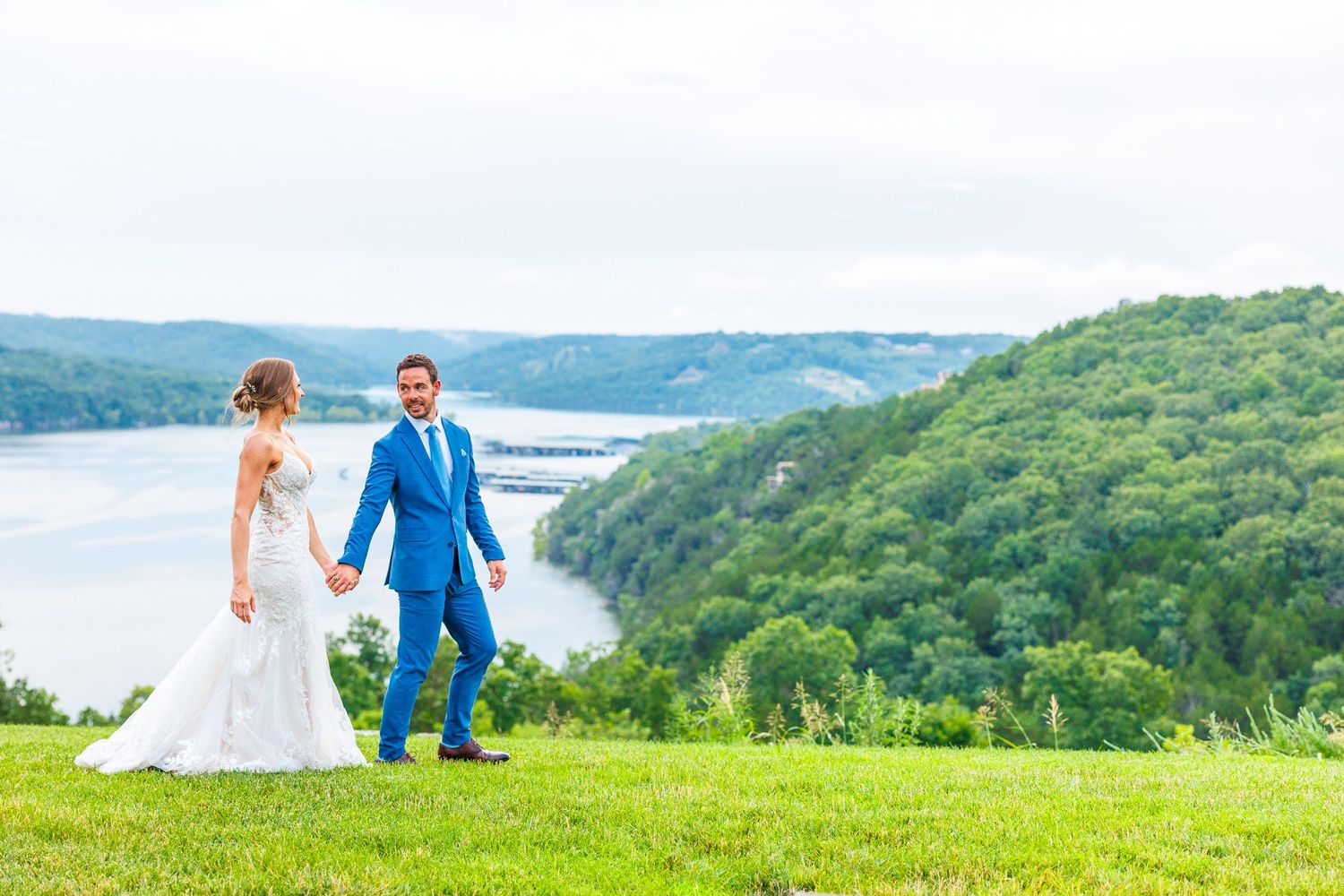 Couple after wedding walks hand in hand through lush green hillside overlooking scenic table rock lake view on wedding day Integrity Hills.