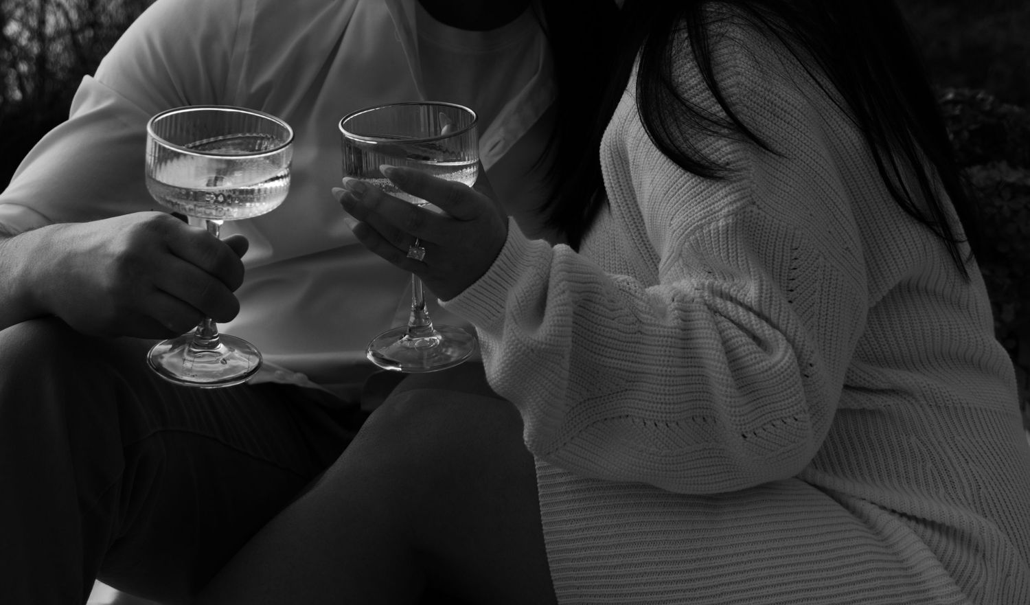 Black and white close-up of couple clinking elegant cocktail glasses together outdoors.