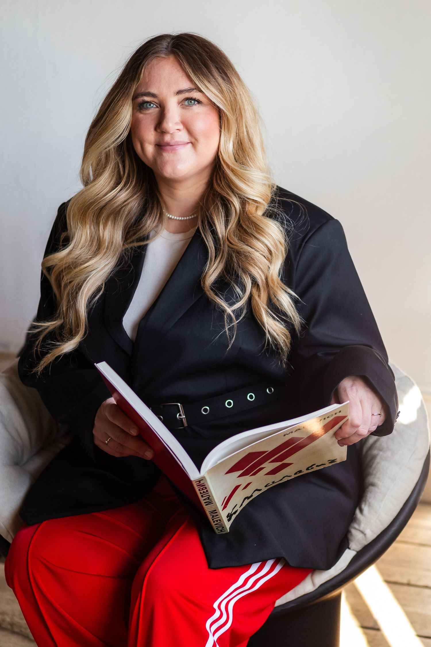 A person in a black blazer and red track pants sits comfortably in a chair while reading a book.