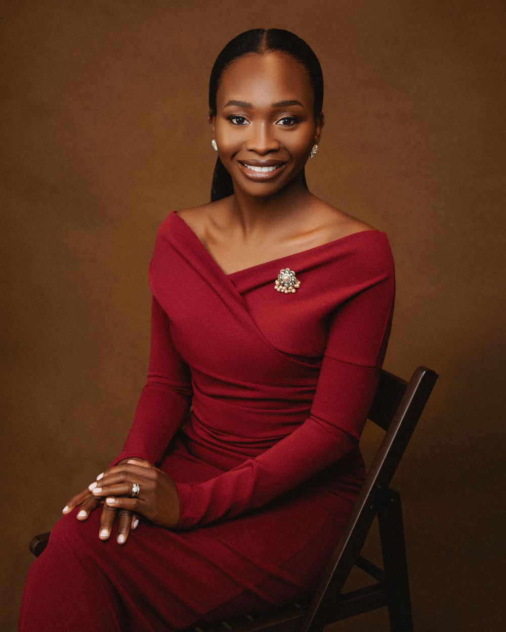 A person in an elegant red off-shoulder dress sitting on a chair against a brown backdrop with a brooch accent.