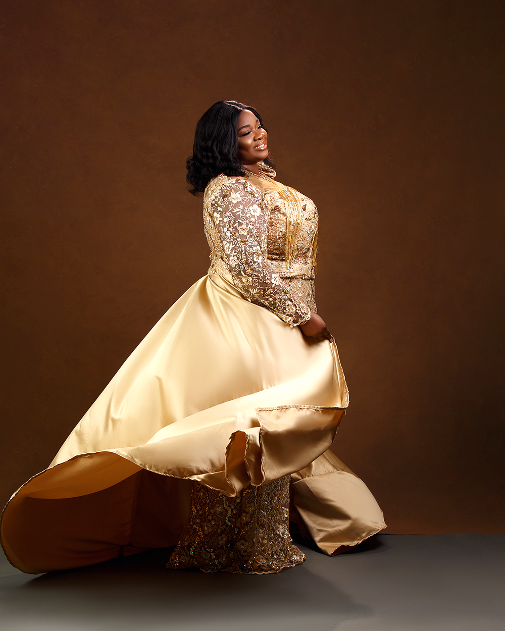 Elegant evening gown in cream satin and lace with flowing skirt photographed against brown backdrop in studio portrait.