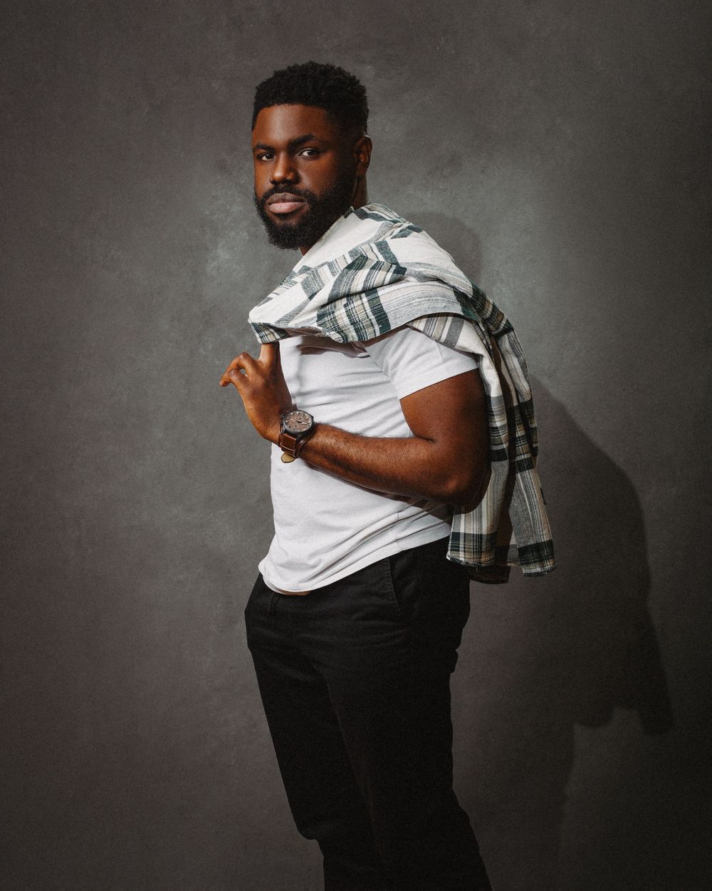 Studio portrait showing person in white t-shirt and black pants carrying plaid jacket over shoulder against gray background.
