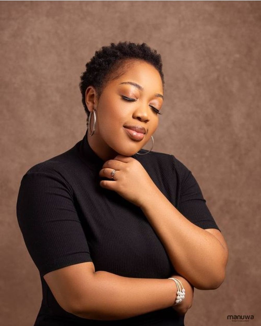 Professional portrait of woman in black top with short hair and elegant pose against brown backdrop.