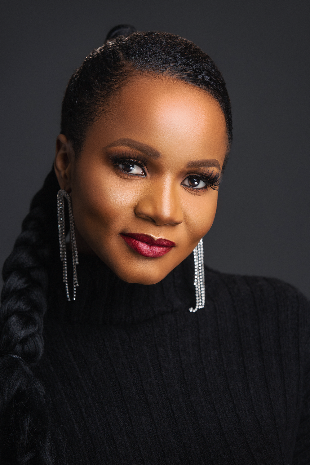 Professional portrait of someone wearing black sweater and sparkly dangle earrings with flawless makeup against dark background.