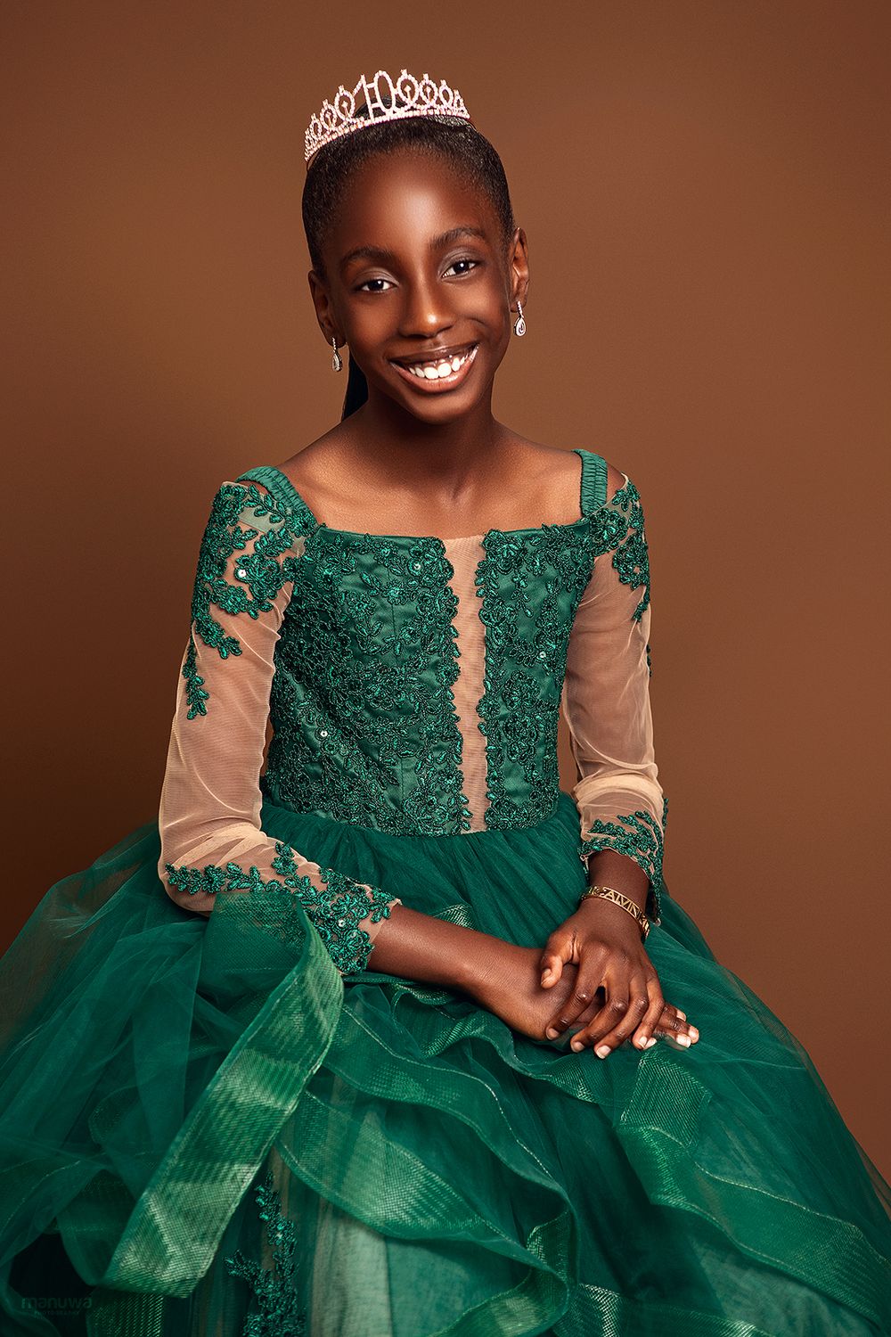 Beautiful formal portrait in emerald green ball gown with lace details and sparkly tiara against brown background.