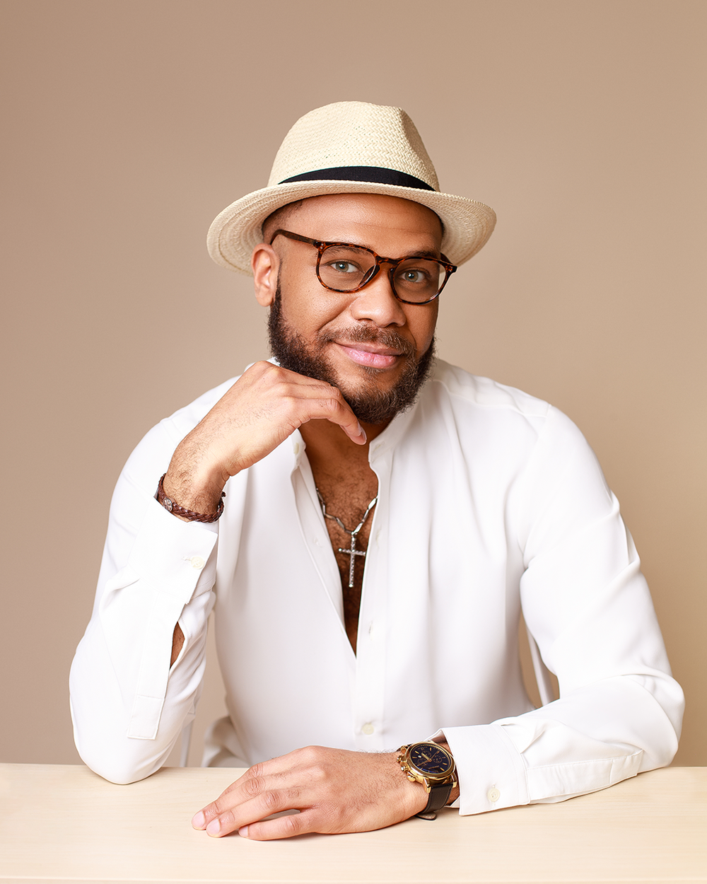 Stylish person in white shirt and fedora smiling while seated at table wearing glasses and watch.