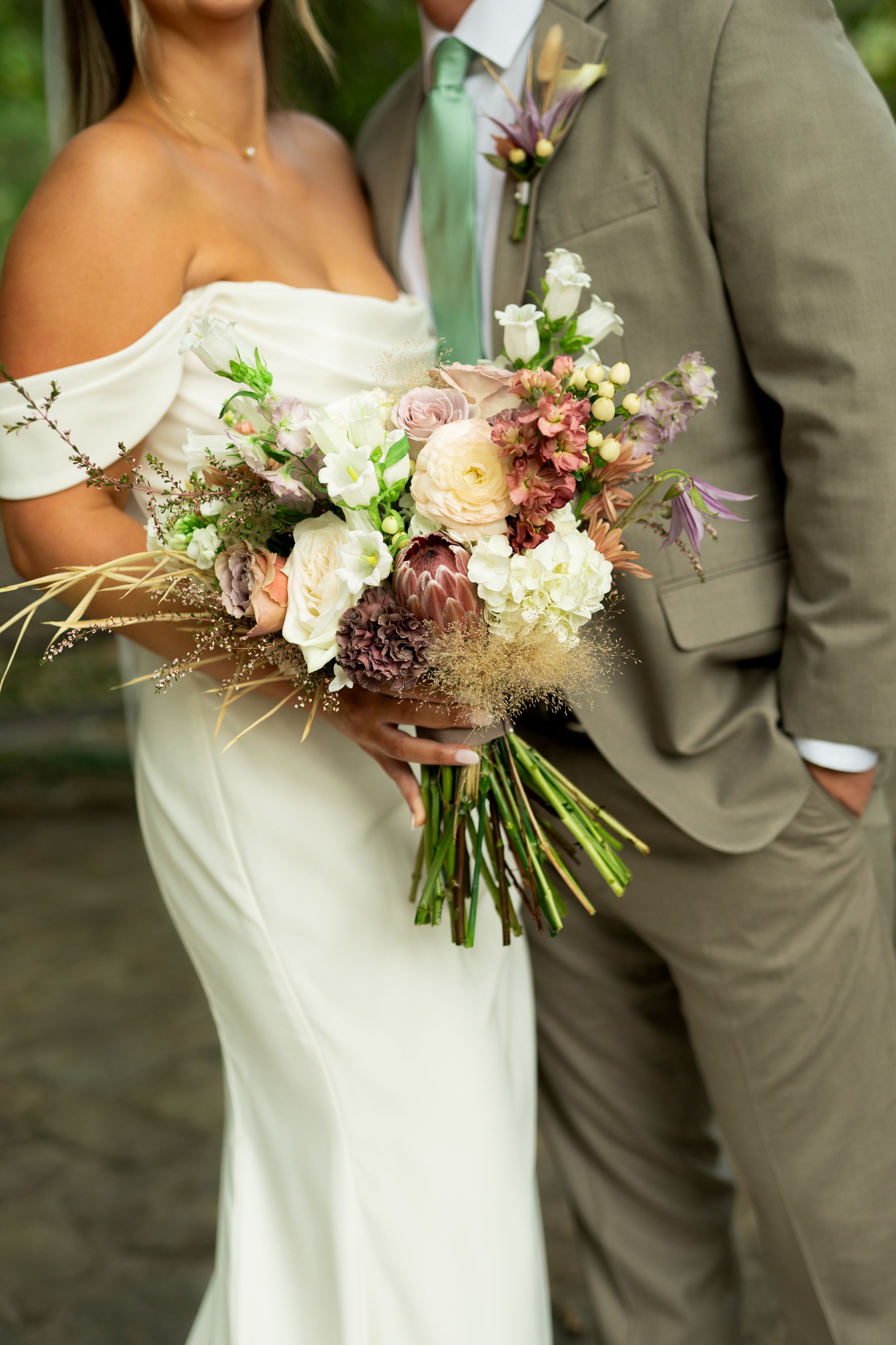 Wedding couple detail shot featuring white bridal gown and grey suit with rustic floral bouquet.