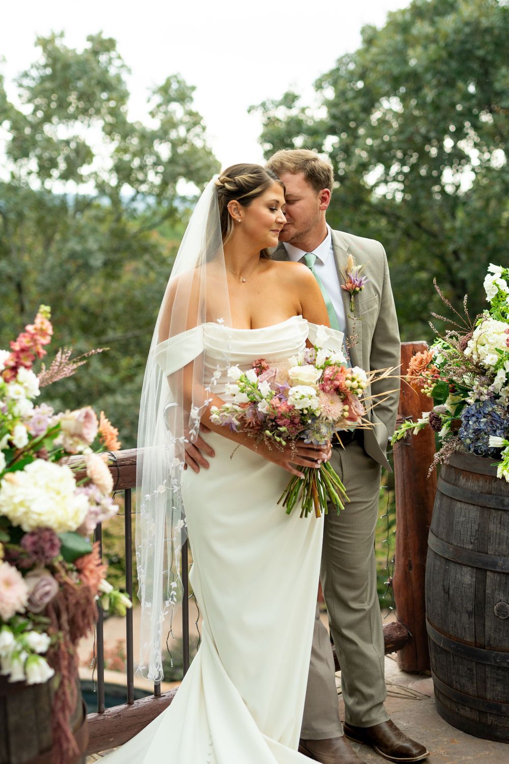 Elegant wedding couple poses near rustic wine barrels decorated with colorful flowers.