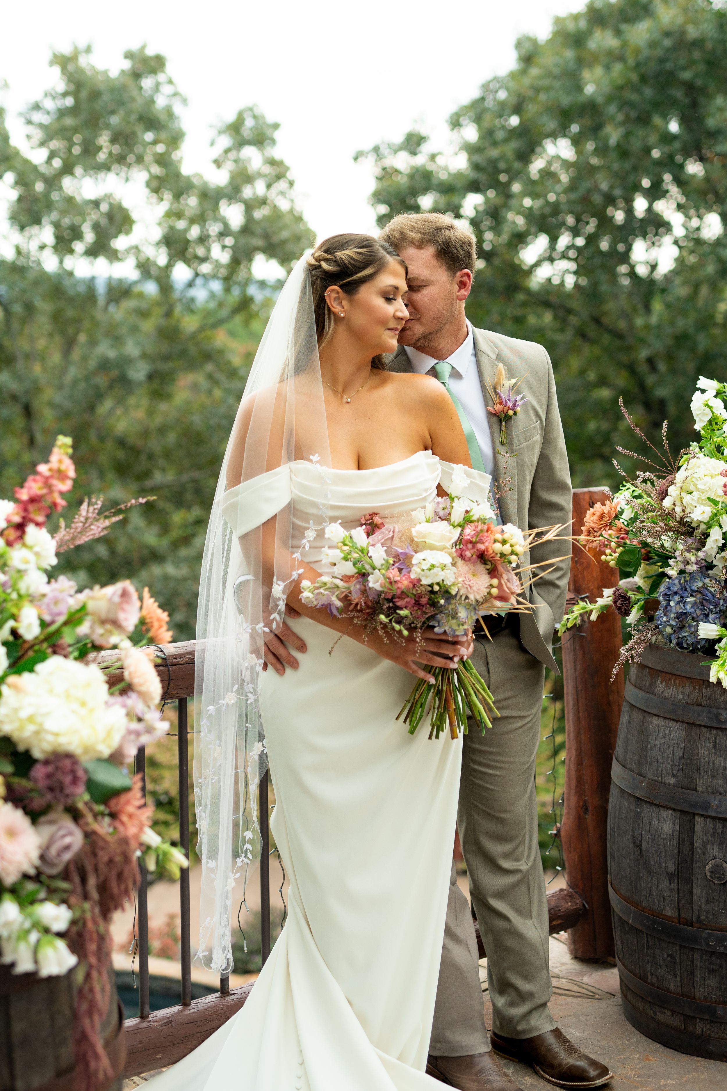 Elegant wedding couple poses near rustic wine barrels decorated with colorful flowers.