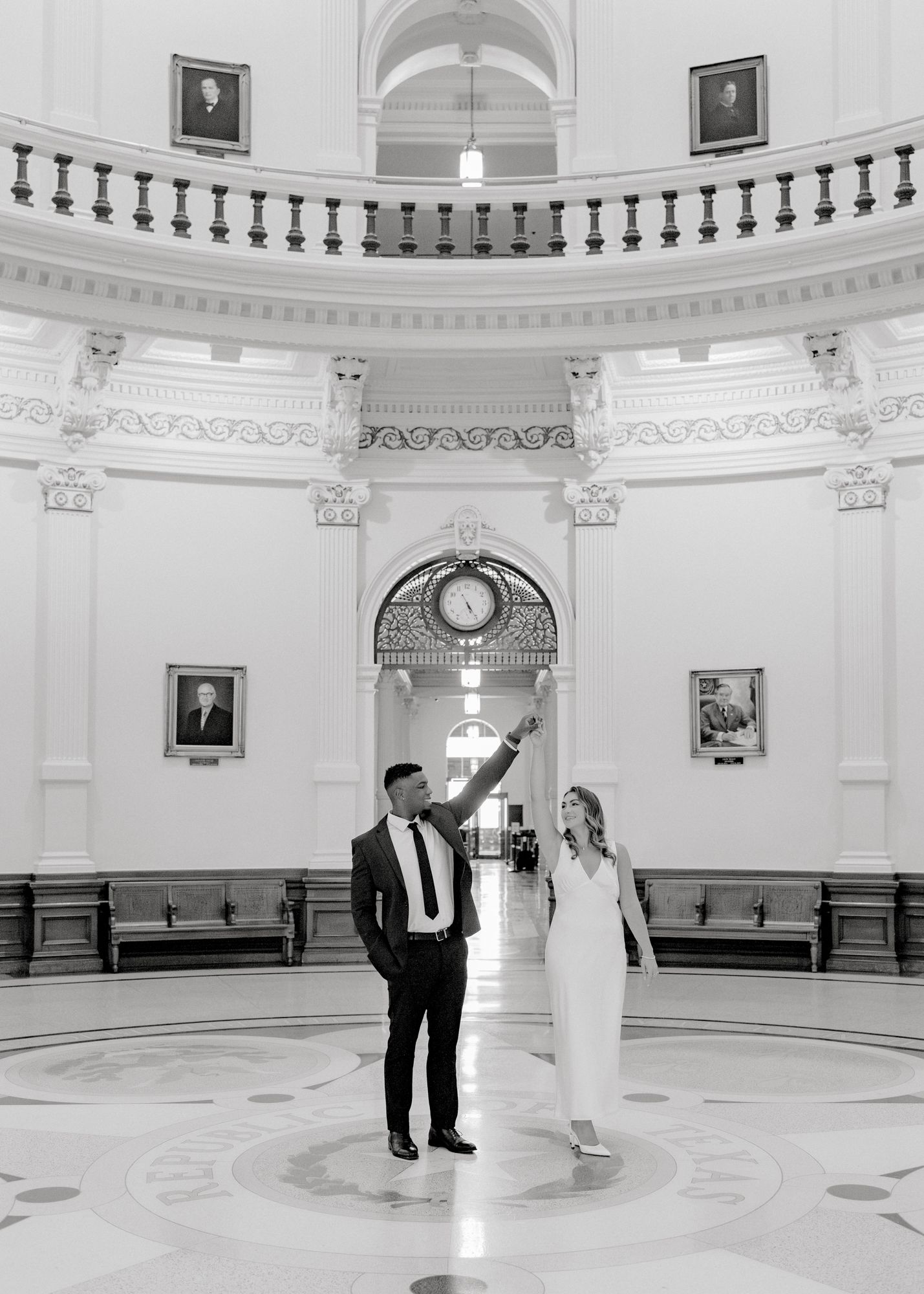 A couple poses in an elegant rotunda with ornate architecture and a black and white marble floor.